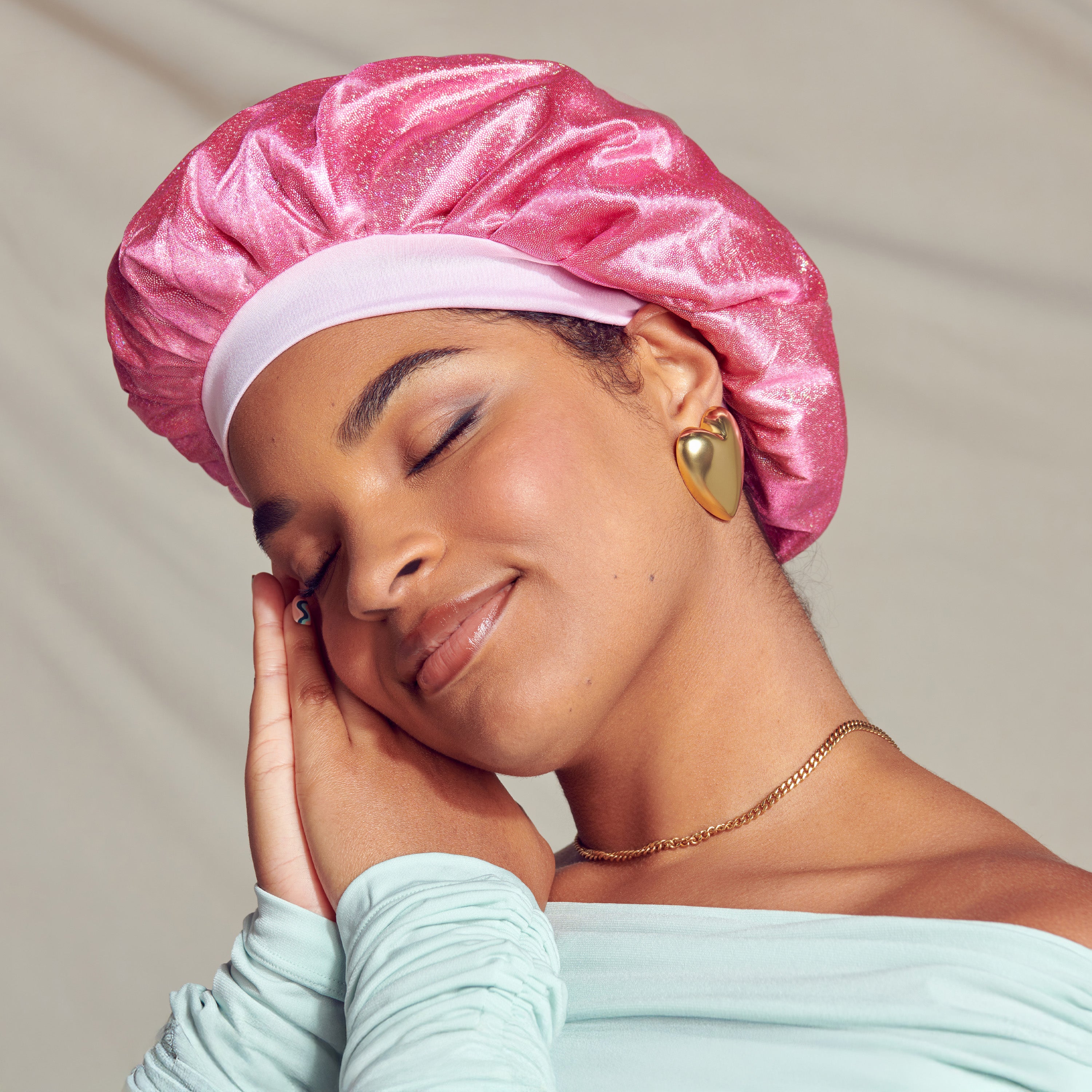 Woman with eyes closed and smiling softly, wearing a shiny pink pearl satin bonnet cap with a light pink band, gold heart-shaped earrings, a gold chain necklace, and a light mint green top.