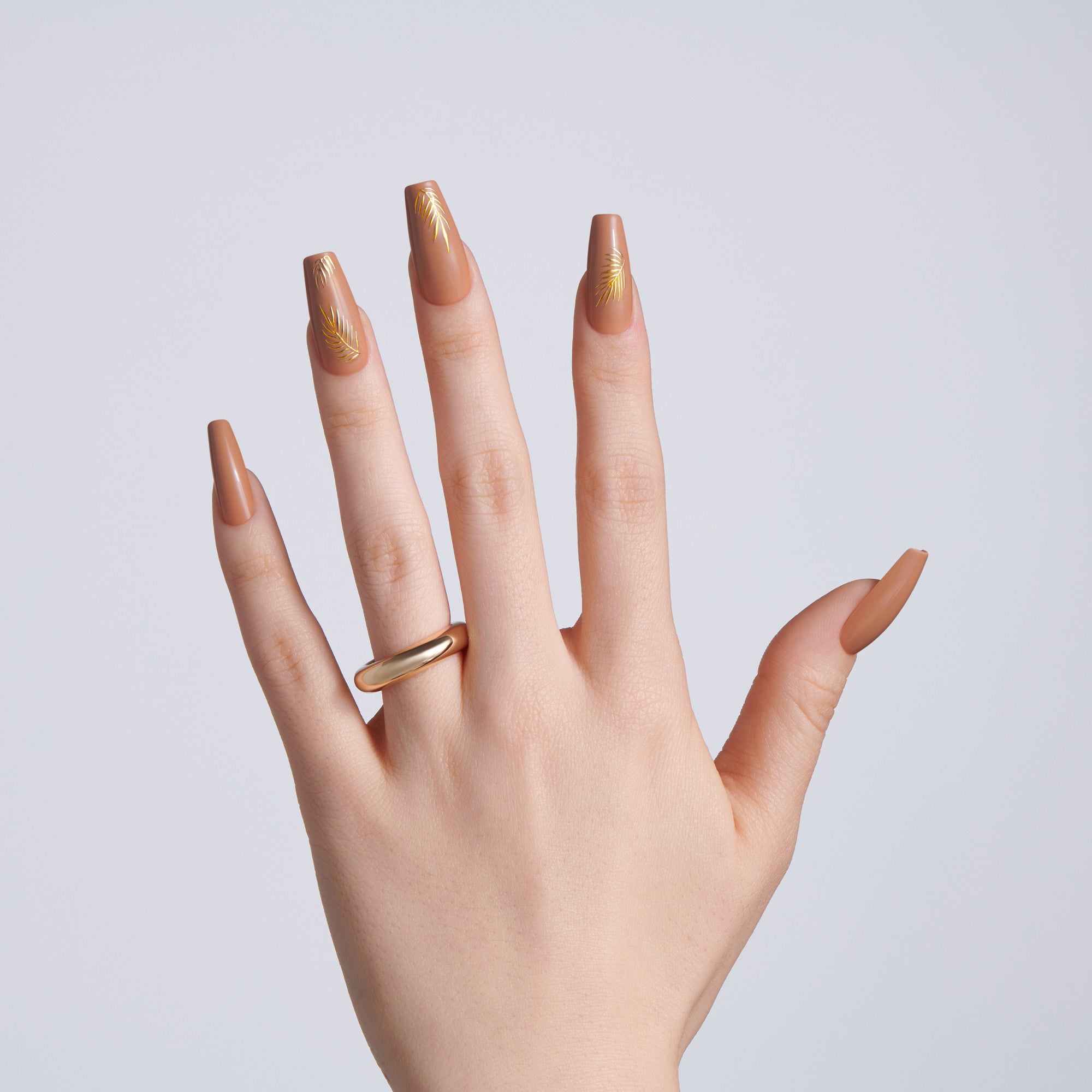 Hand wearing a gold ring with long coffin-shaped nude brown nails, some featuring gold leaf accent designs, against a light gray background