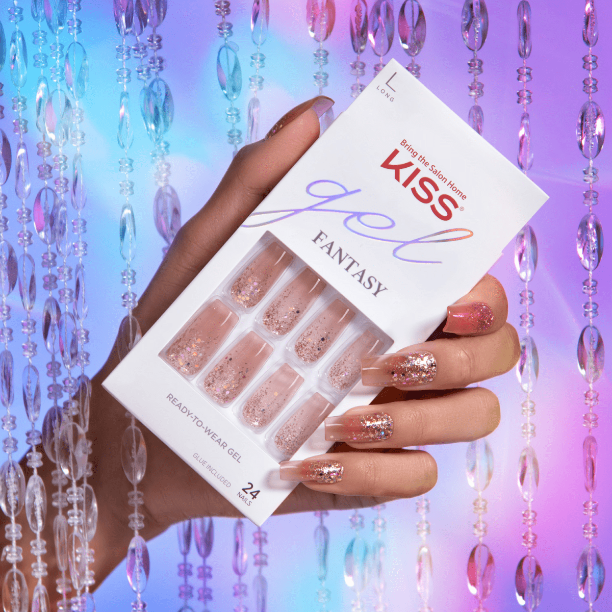 Hand holding KISS Gel Fantasy Nails packaging with long, glittery pink gel nails matching the 24 ready-to-wear nails shown in the package against a colorful beaded curtain background.