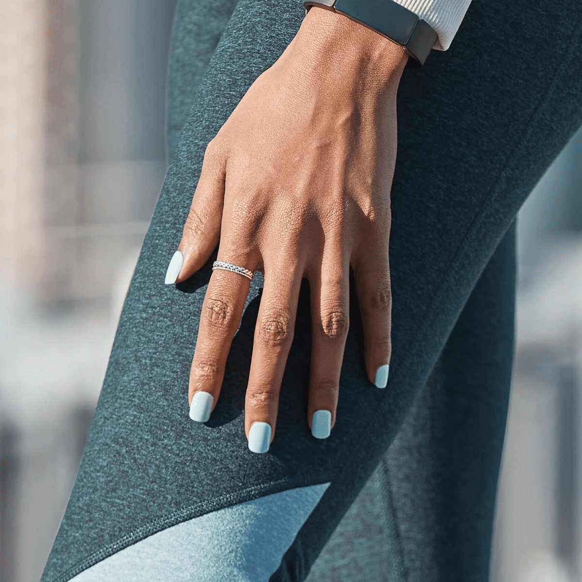 Hand with medium skin tone wearing pale blue square-shaped press-on nails, resting on dark teal athletic leggings, showcasing the imPRESS Rain Check manicure.