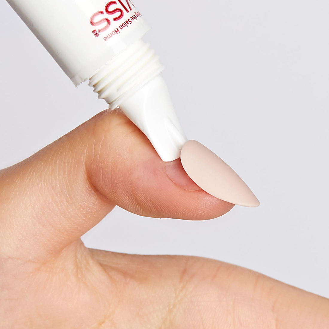 Close-up of a hand applying a white tube of KISS Glue OFF remover to detach a blush pink press-on nail from the thumb.