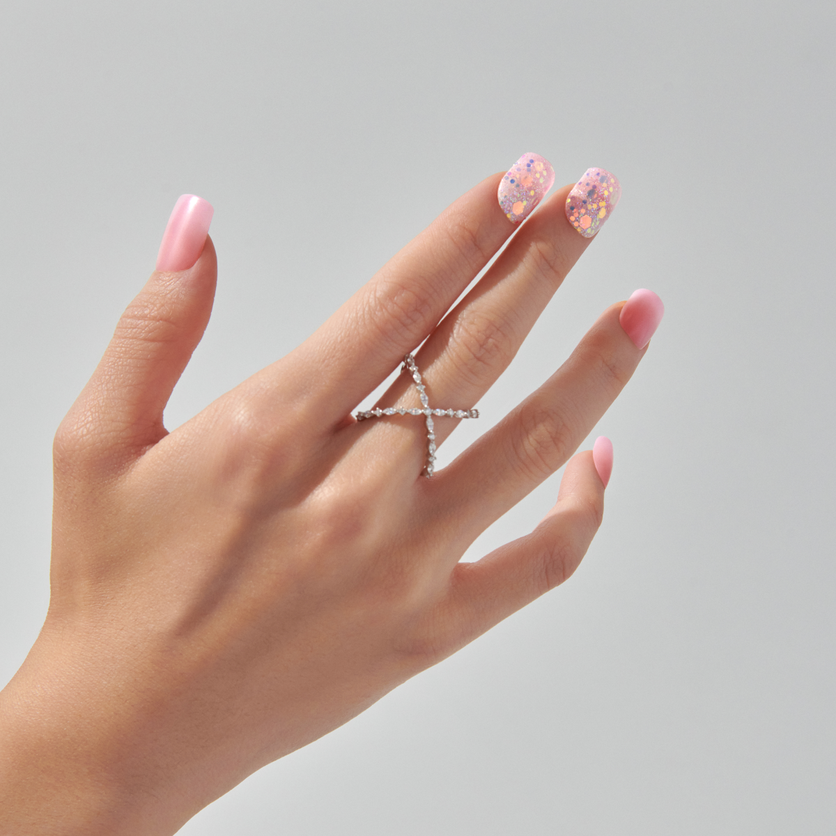 Hand with wide fit press-on nails featuring a glossy pink base and two nails with multicolored glitter dots, wearing a silver crisscross ring.