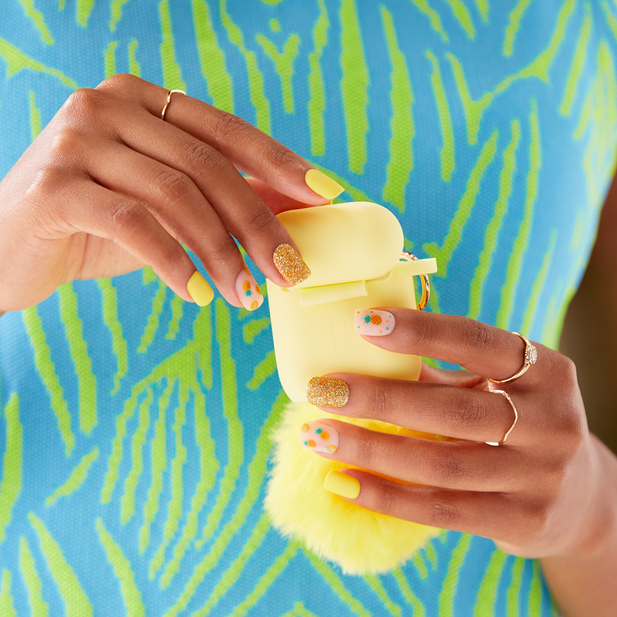 Hands with bright yellow matte, gold glitter, and pineapple-patterned press-on nails holding a matching yellow AirPods case with a fluffy keychain, against a blue and green patterned top.
