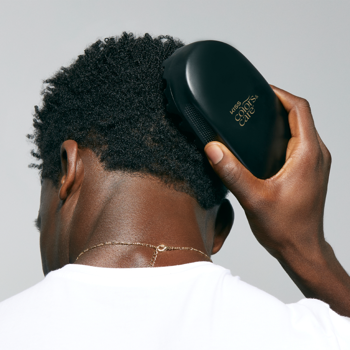 Man with short curly black hair wearing a white shirt, using a black KISS Colors & Care hair brush on the back of his head.