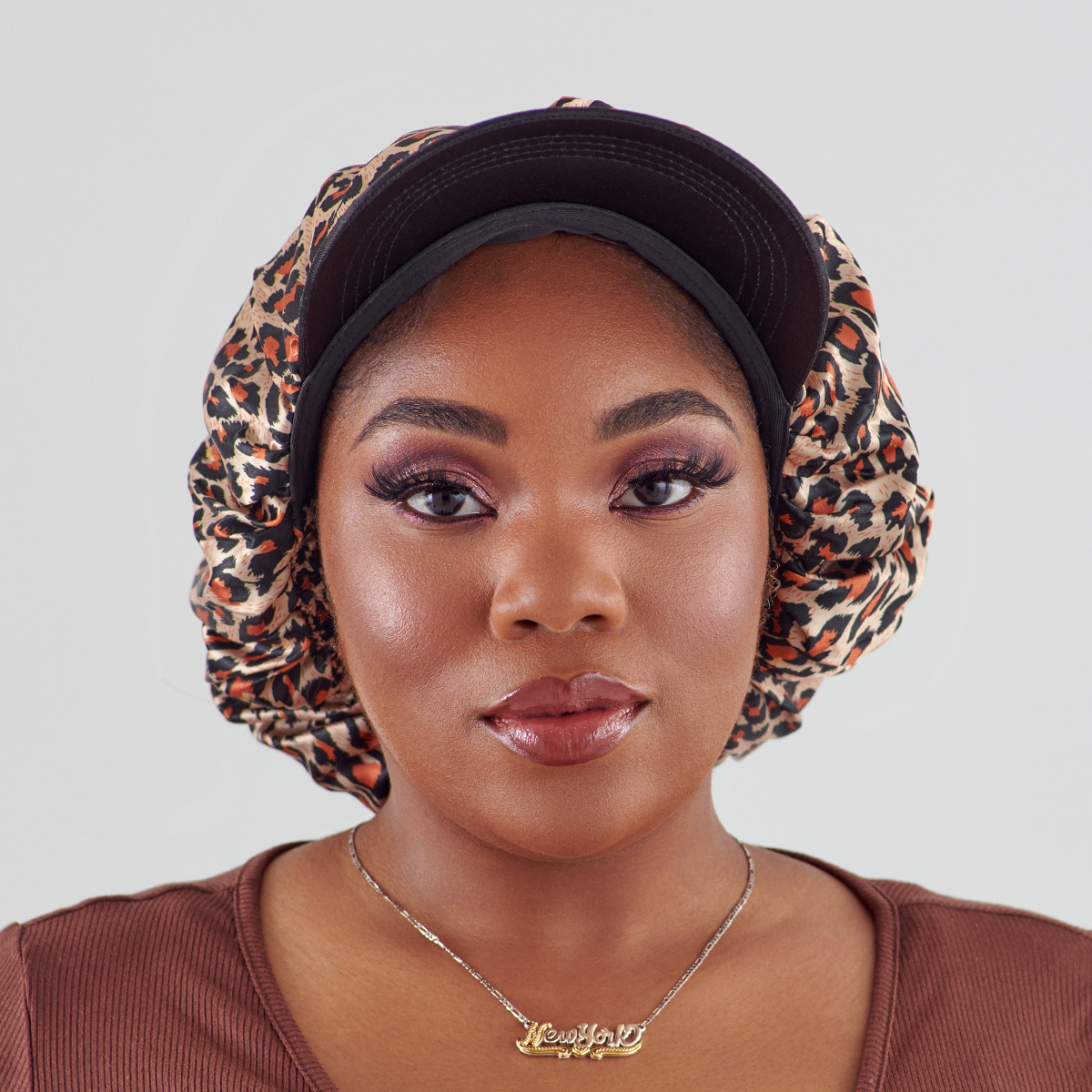 Woman facing forward wearing a silky satin bonnet cap with a black visor and leopard print fabric gathered around the head, styled for protective hair care.
