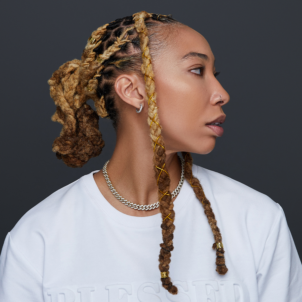 Profile of a person with blonde and brown braided hair styled with gold metal braid charms and wire spirals, wearing a white shirt and silver chain necklace.