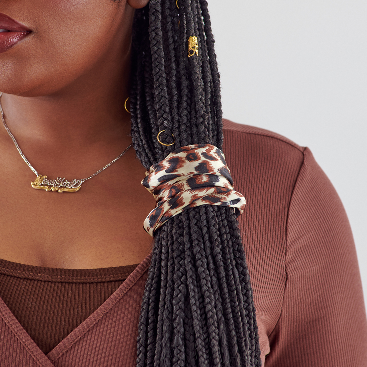 Black and brown leopard print silky satin scrunchie wrapped around thick black braided hair, worn by a person in a ribbed brown top.