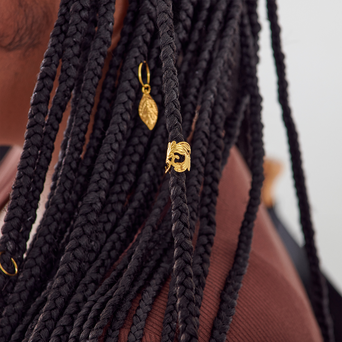 Close-up of black braided hair adorned with two gold-colored braid charms, including a leaf-shaped charm and a decorative ring charm.