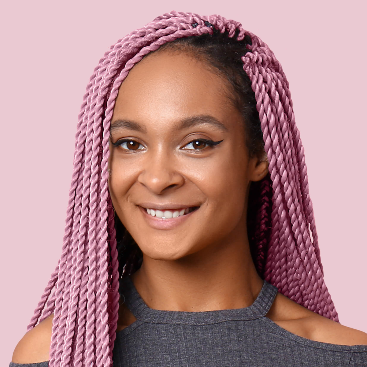 Woman with long, twisted rose-colored braids, wearing a dark gray cold-shoulder top, smiling against a light pink background.