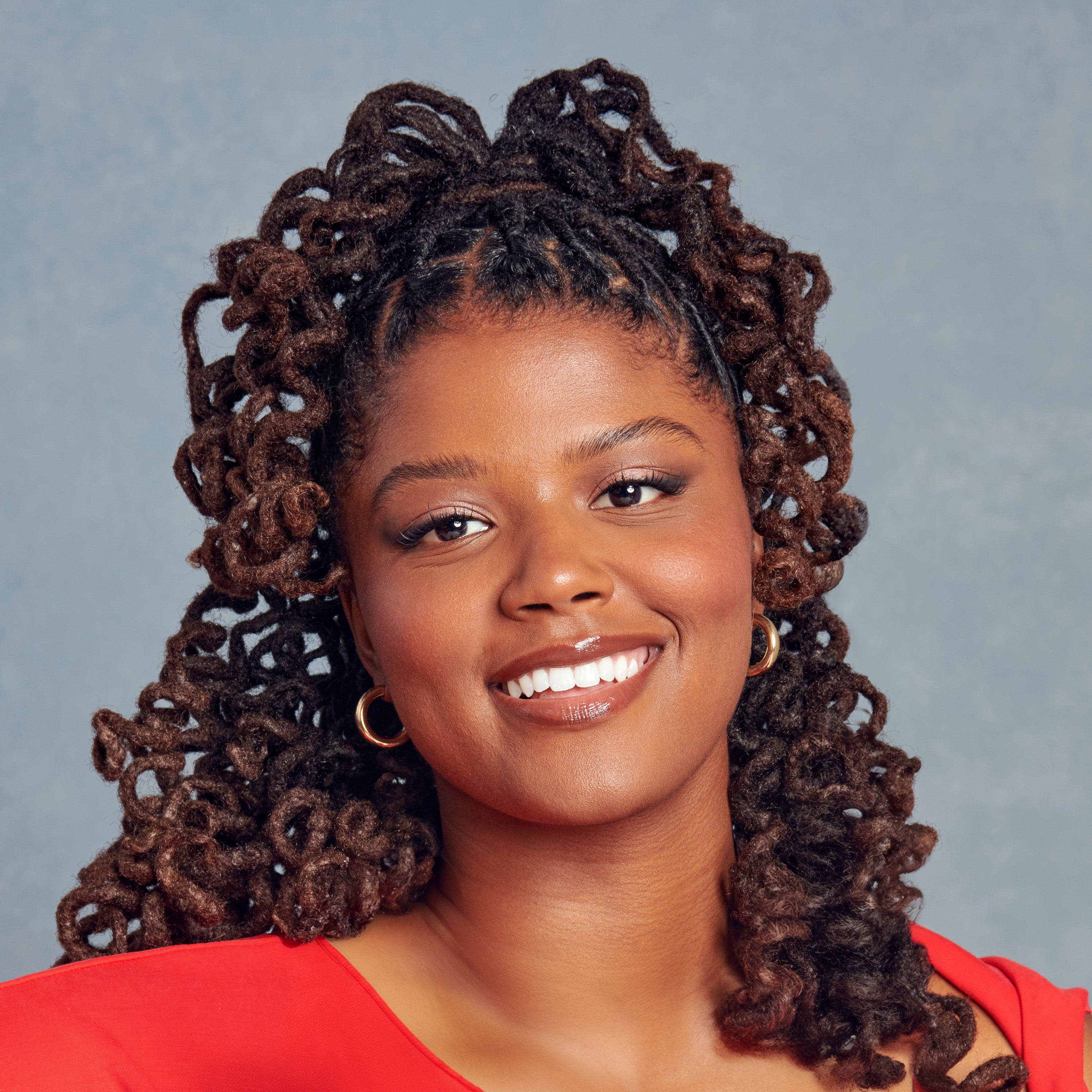 A close-up of a smiling woman with styled curly hair wearing gold hoop earrings and a red top, against a neutral gray background.