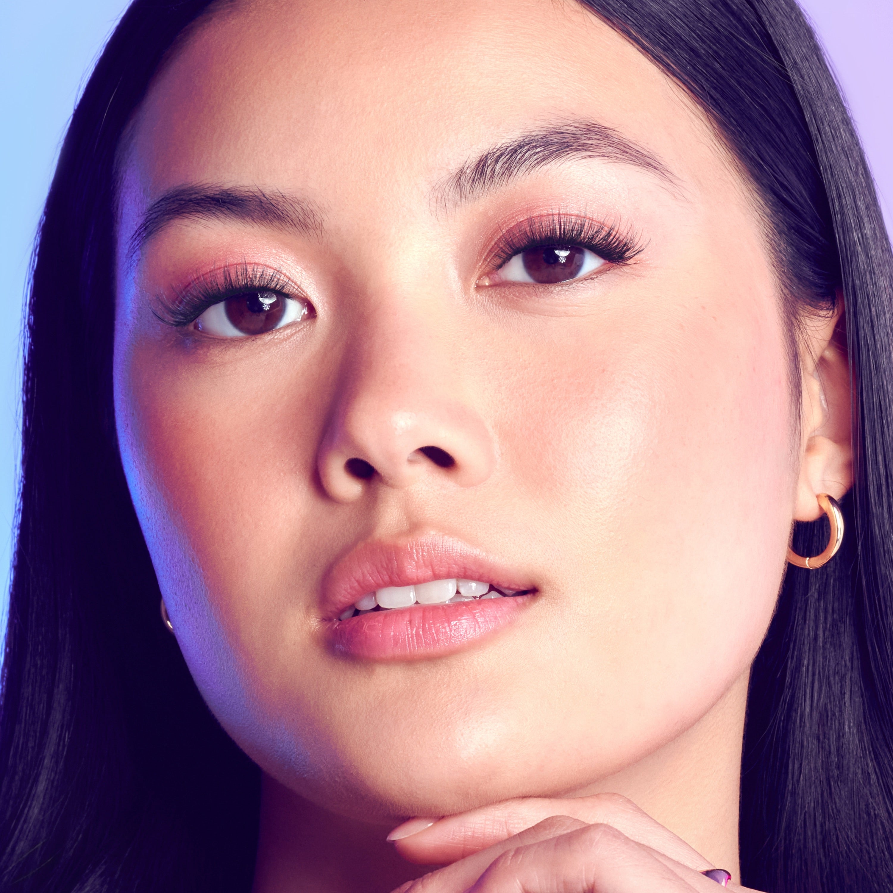 Close-up of a woman with dark brown eyes and long, voluminous lashes, wearing gold hoop earrings and natural pink lip color, showcasing eyelash extensions.