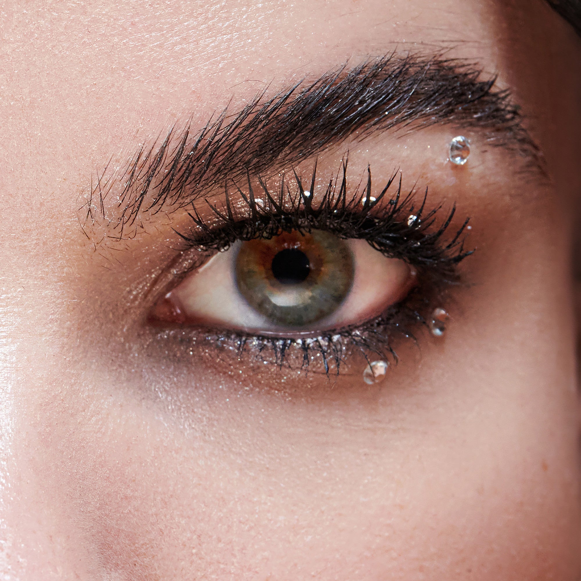 The image features a close-up of a human eye, showcasing striking details. The eye has a vibrant, multi-colored iris with shades of green and brown. Long, dark eyelashes frame the eye, enhancing its allure. A small, sparkling droplet of water or a decorative gem is positioned just below the eye, adding a touch of glamour. The skin around the eye appears smooth, with a subtle shimmer, contributing to an overall captivating and artistic look.