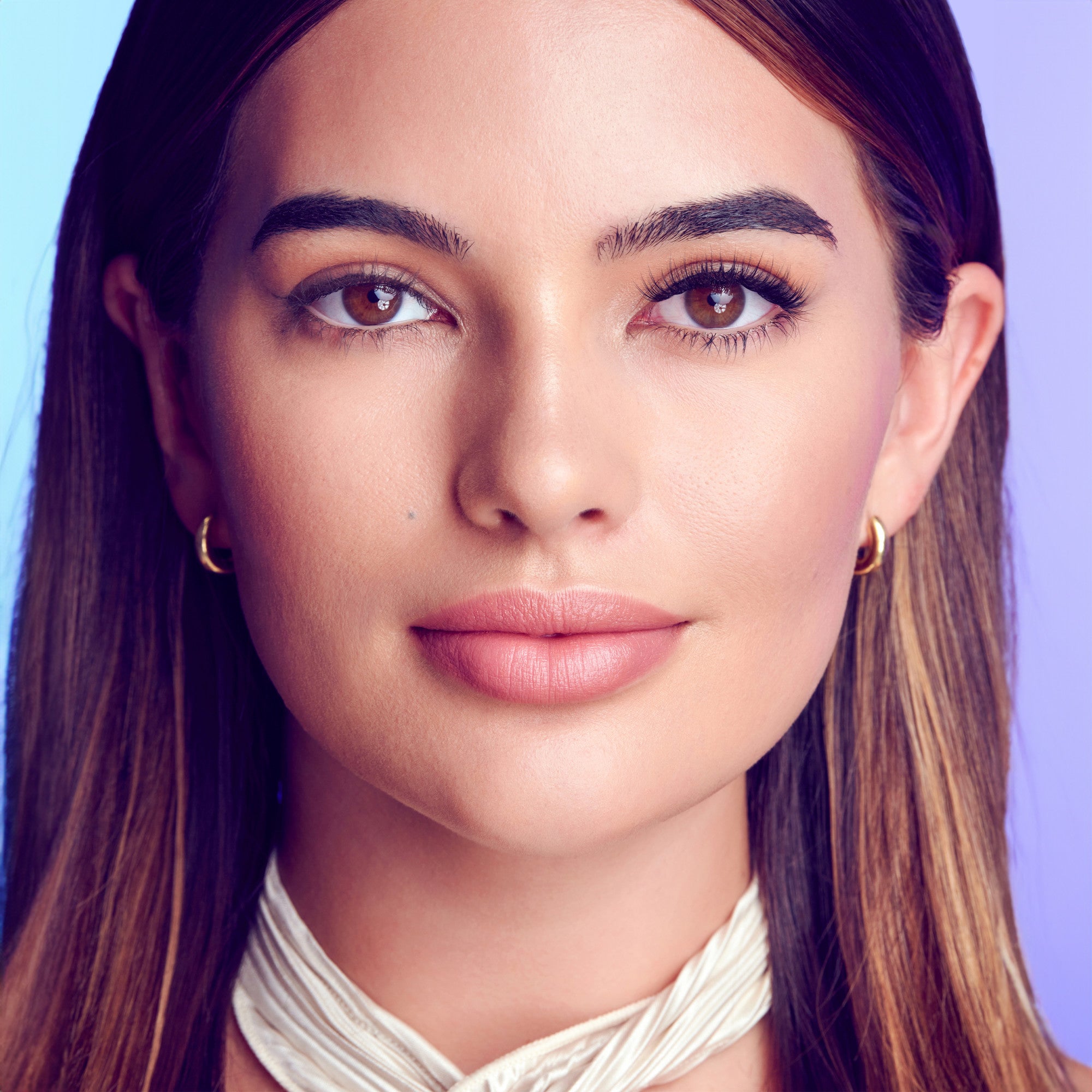 The image features a close-up portrait of a young woman with long, straight hair that has a blend of dark and light tones. She has a smooth complexion and well-defined features, including full lips and expressive eyes. The background is a gradient of soft colors, enhancing the overall aesthetic. The woman is wearing a simple, elegant accessory around her neck, adding a touch of sophistication to her look. Her expression is calm and confident, making the image striking and engaging.