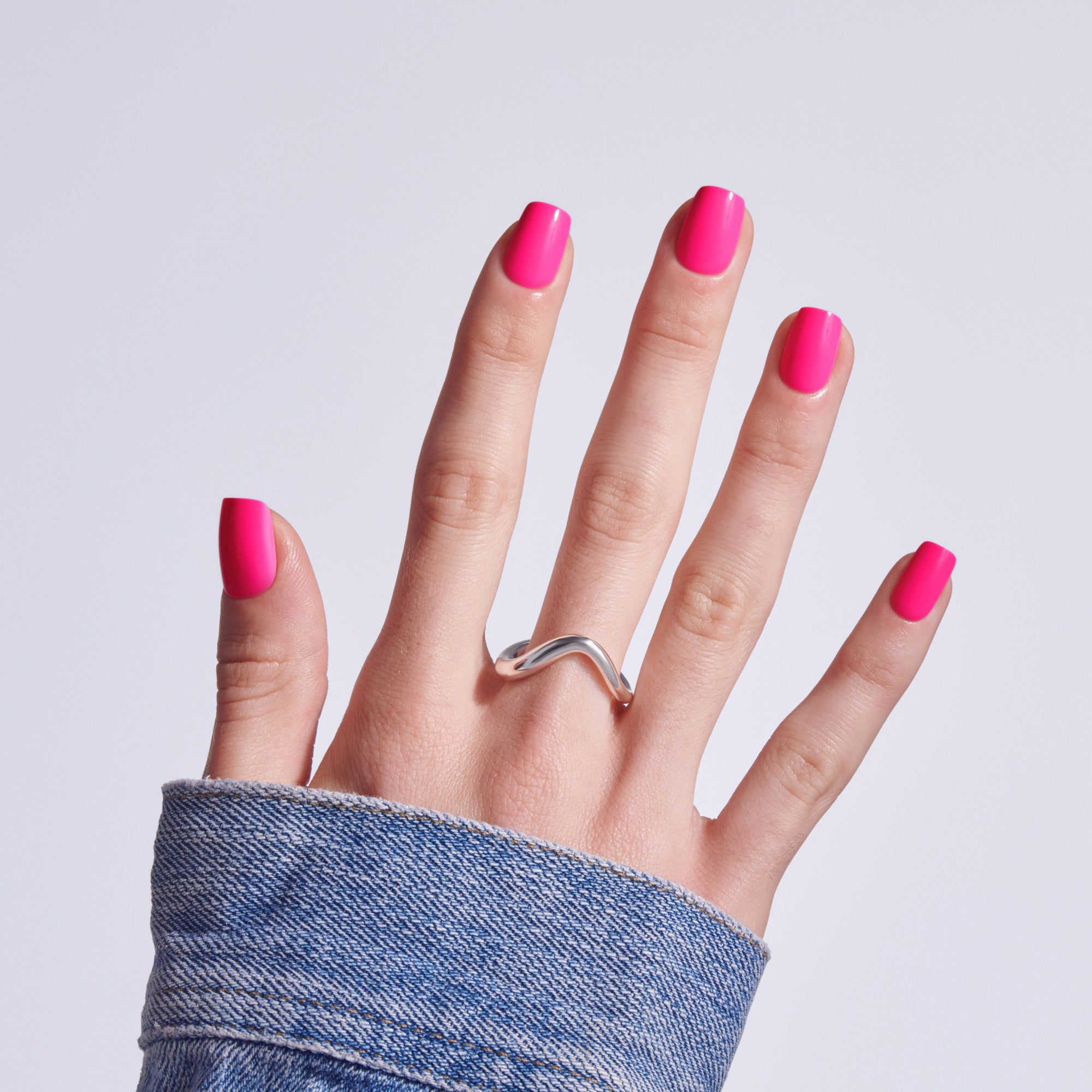Hand displaying bright pink nail polish with silver curved ring against white background.
