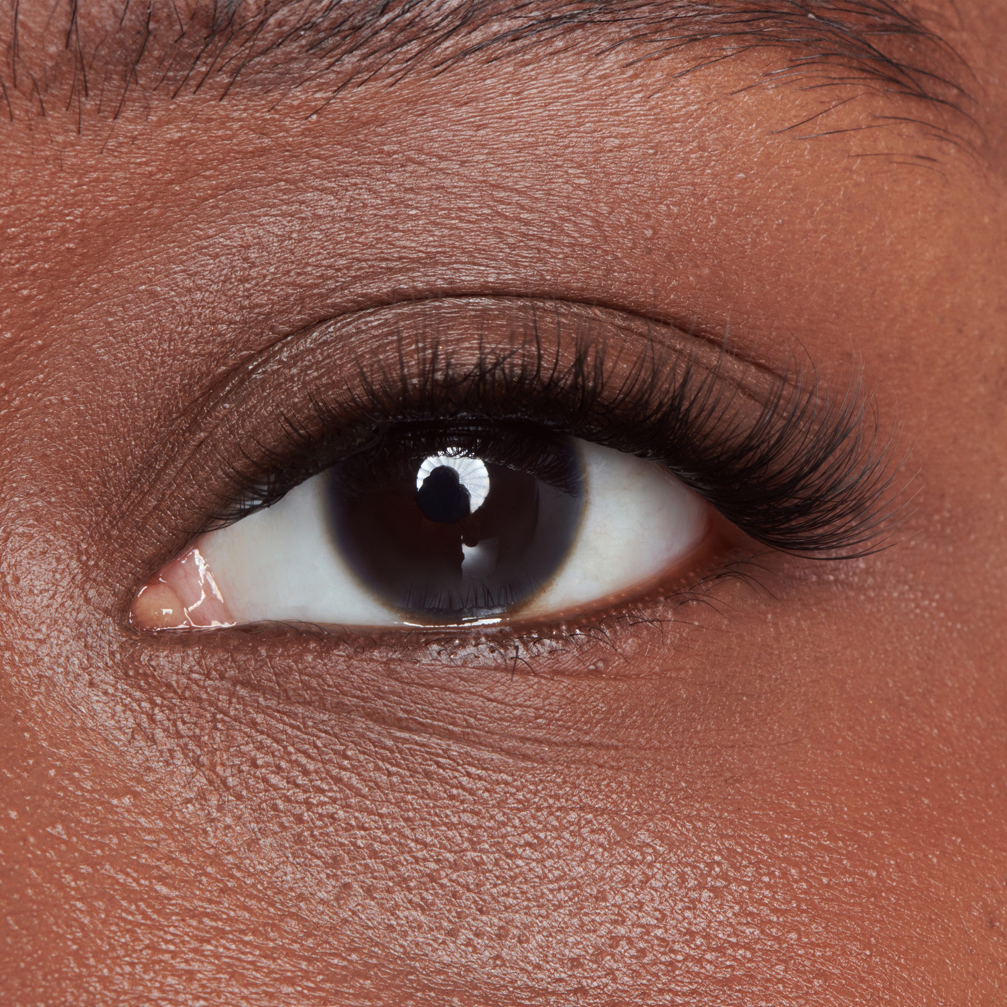 The image features a close-up of a human eye, showcasing a rich, dark brown iris and a well-defined pupil. The skin surrounding the eye is smooth and has a warm tone, highlighting the natural texture. Long, dark eyelashes frame the eye, adding to its expressive appearance. The overall focus is on the eye itself, emphasizing its depth and detail.