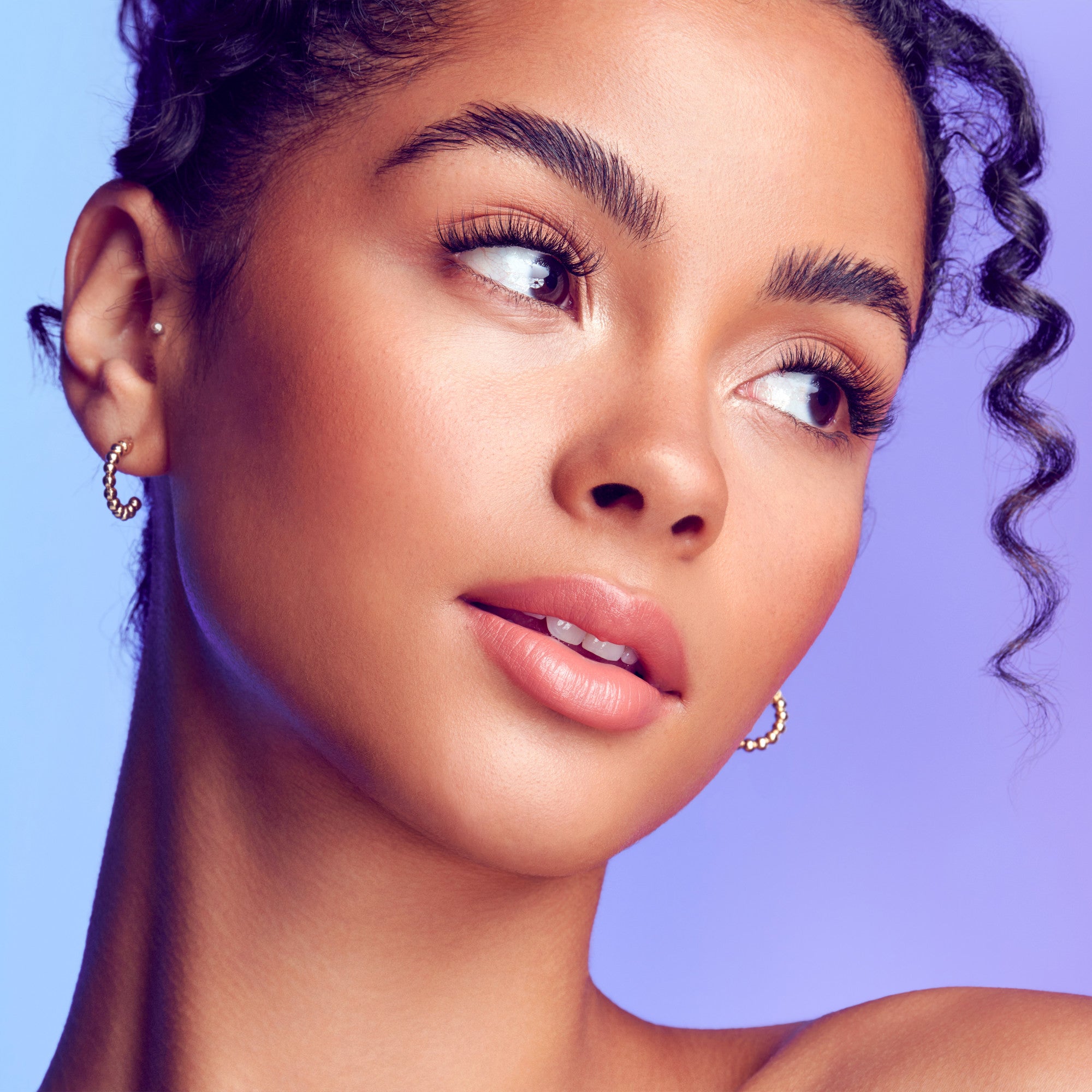 The image features a close-up portrait of a young woman with a radiant complexion. She has curly hair styled elegantly, with some strands framing her face. Her makeup is subtle yet enhances her natural beauty, highlighting her expressive eyes and full lips. She wears small hoop earrings that add a touch of sophistication. The background is a gradient of soft purple tones, creating a dreamy and vibrant atmosphere.