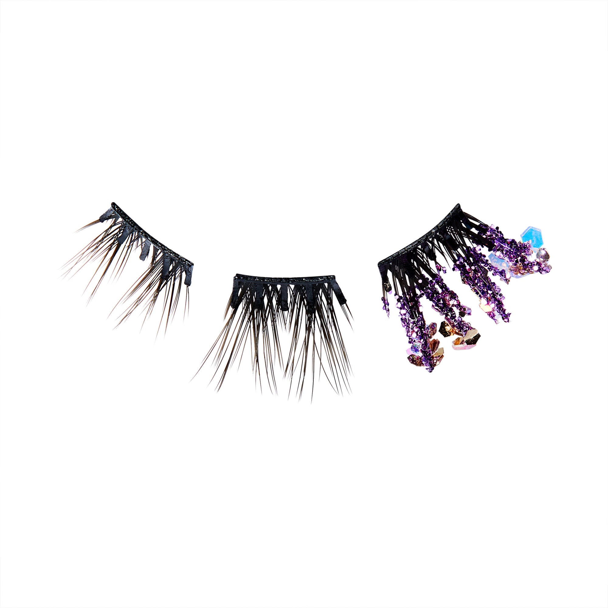 The image features a set of dramatic false eyelashes. The lashes are predominantly black, with varying lengths and styles. One section has a more traditional lash design, while the other two sections are adorned with decorative elements, including purple crystals and beads that hang down, adding a unique and glamorous touch. The overall look is bold and eye-catching, perfect for a statement makeup look.
