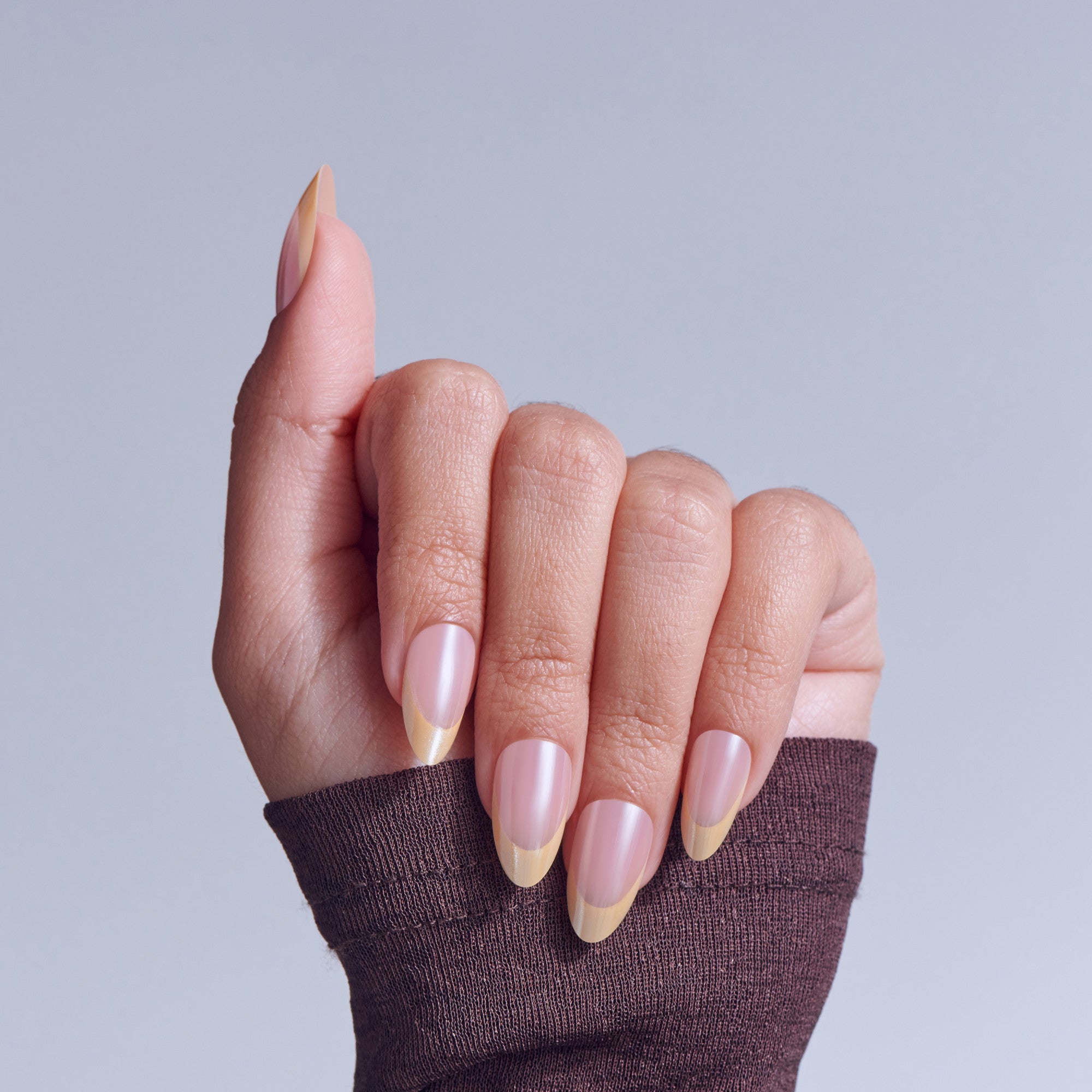 Hand displaying almond nails with pink polish and nude/gold ombre tips against gray background.