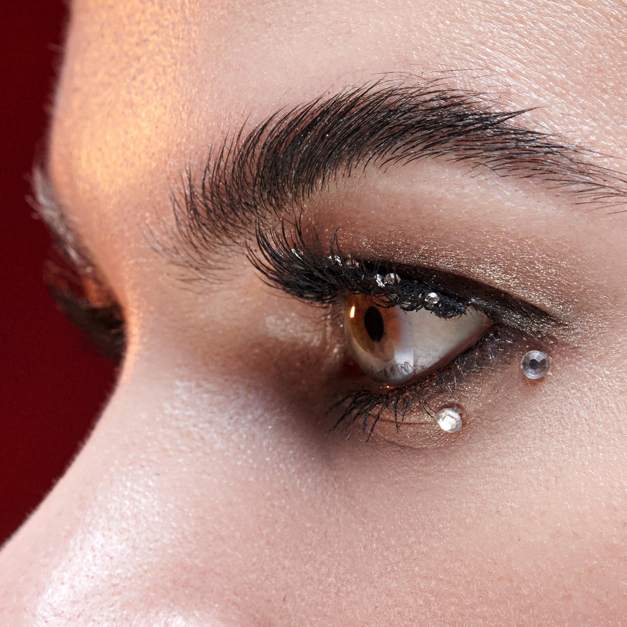 The image features a close-up of an eye, showcasing intricate details. The eye has a striking golden-brown iris, accentuated by long, dark eyelashes. The eyelid is adorned with a subtle shimmer, enhancing the overall look. Additionally, there are decorative elements, including small gemstones placed near the eye, adding a touch of glamour. The background appears to be a soft, blurred red, which contrasts beautifully with the eye&