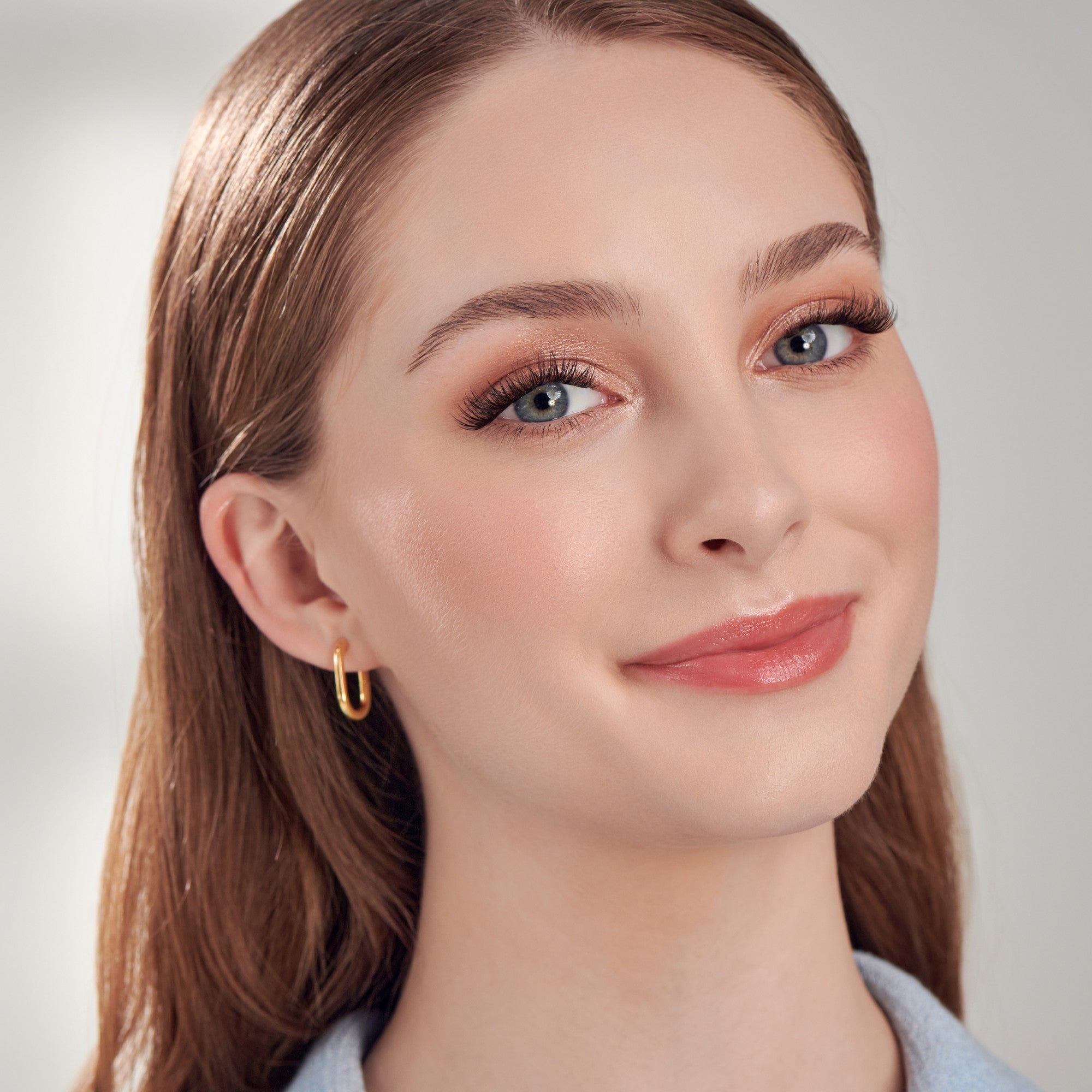 The image features a young woman with long, straight hair and a warm smile. She has a natural makeup look, highlighting her eyes with subtle eyeshadow and long eyelashes. Her lips are painted in a soft, peachy shade. She is wearing a pair of simple gold hoop earrings, which add a touch of elegance to her appearance. The background is softly blurred, creating a clean and professional look.