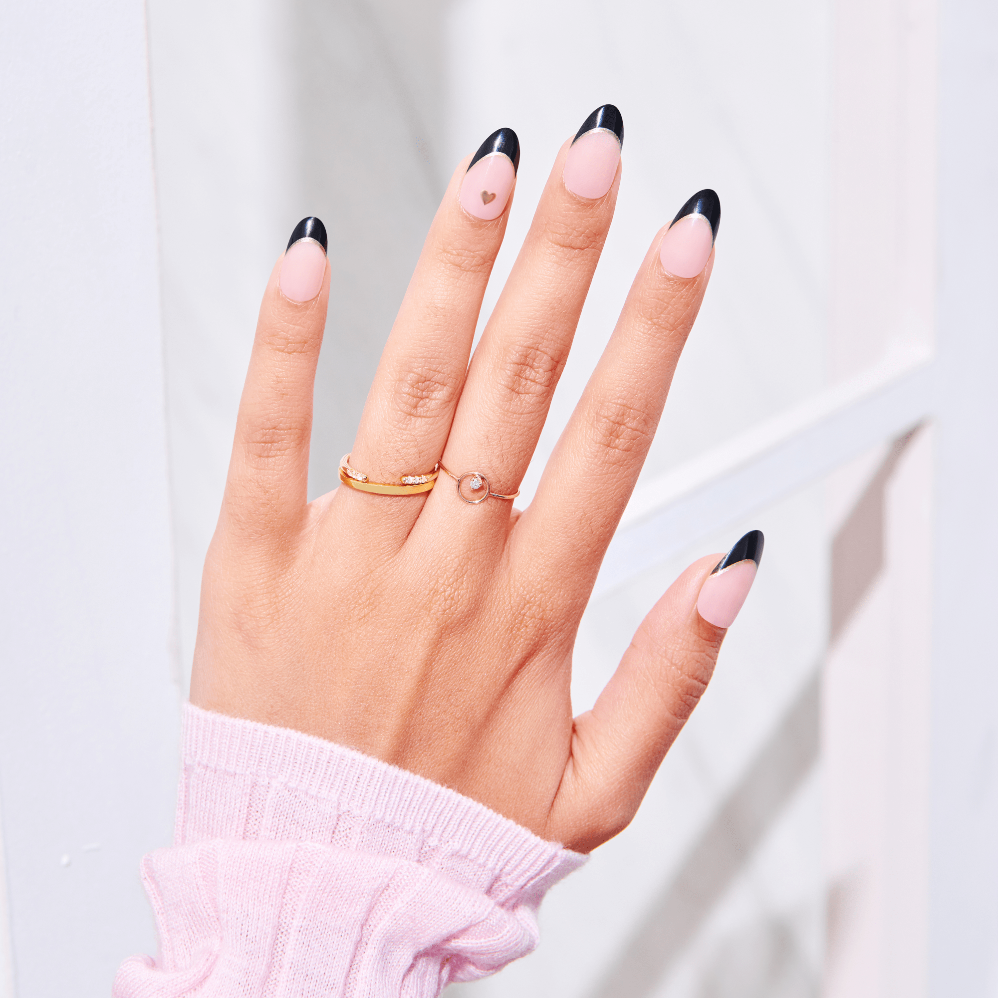 Hand with press-on nails showing a pale pink base and shiny black French tips, with a small gold heart accent on the ring finger. The hand wears two gold rings and a pink sweater sleeve.