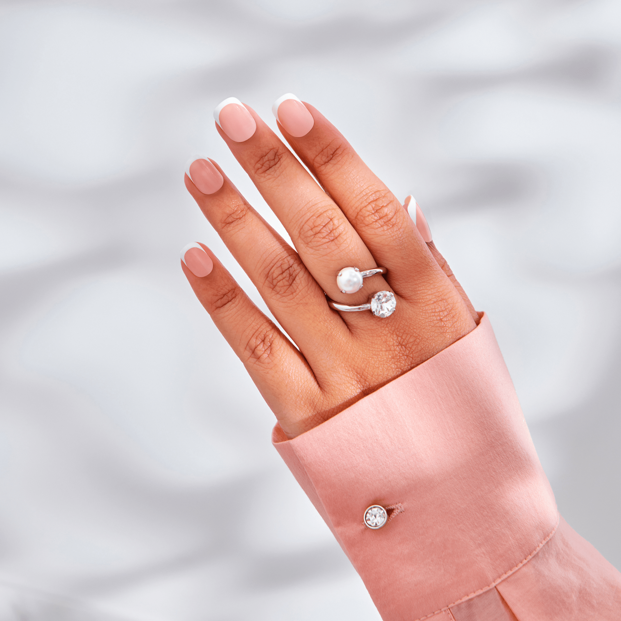 Hand with light pink French press-on nails featuring classic white tips, wearing silver rings with pearl and crystal accents, against a soft light background.