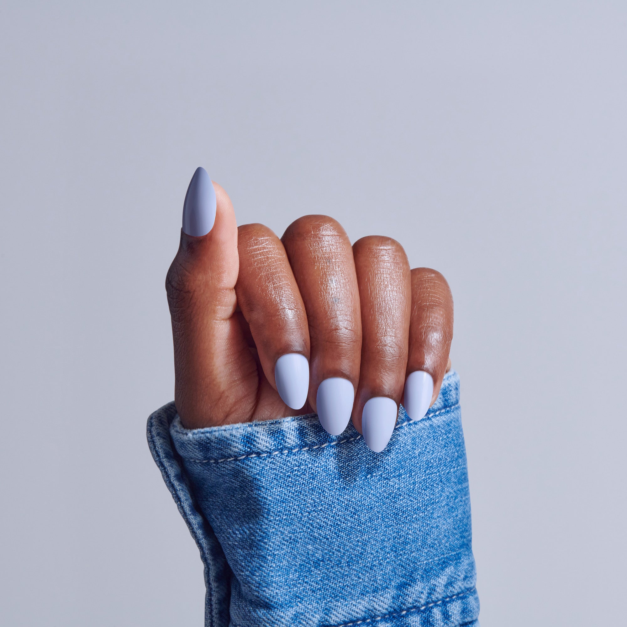 Hand with white almond-shaped manicure emerging from blue denim sleeve against gray background.