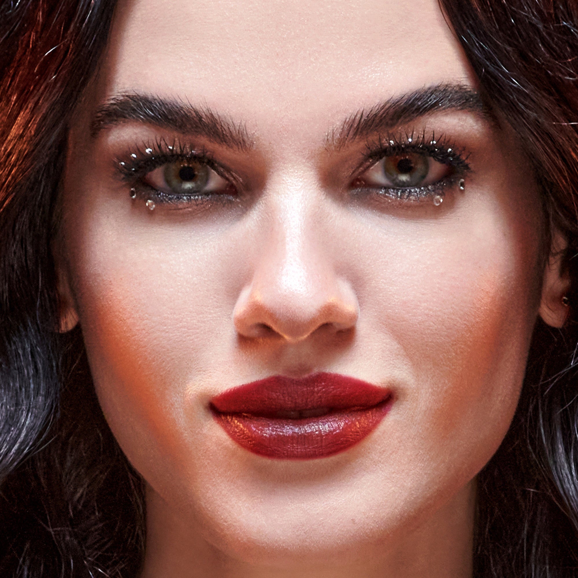 The image features a close-up portrait of a person with striking features. They have long, dark hair that frames their face. Their makeup is bold, with dramatic eye makeup that includes sparkling accents around the eyes and a deep red lip color. The skin has a smooth, radiant quality, enhancing the overall glamorous appearance. The expression is confident and captivating, drawing attention to the eyes and lips.