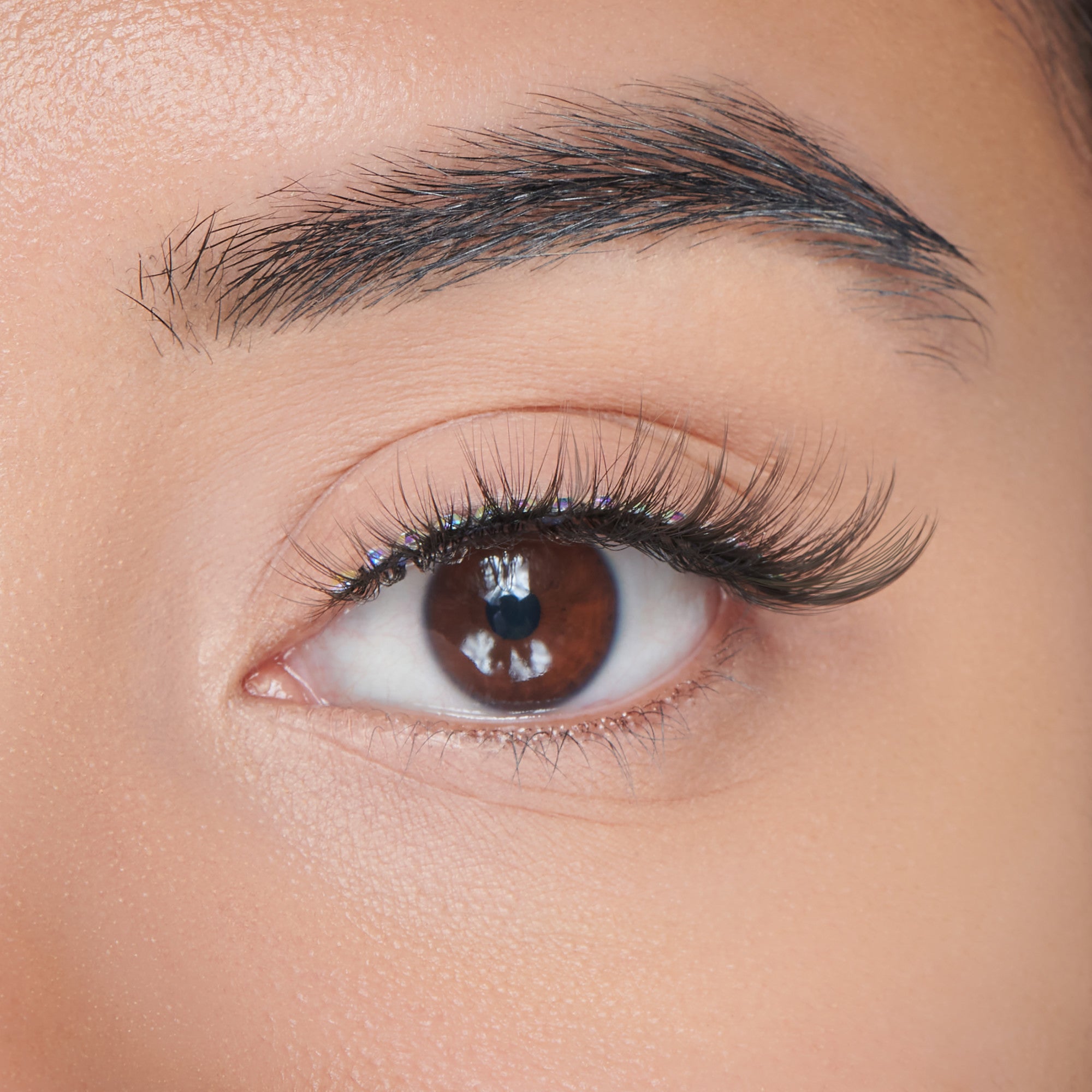 Close-up view of a brown eye wearing long, black false eyelashes decorated with small, iridescent gem stickers for a lustrous shine effect.