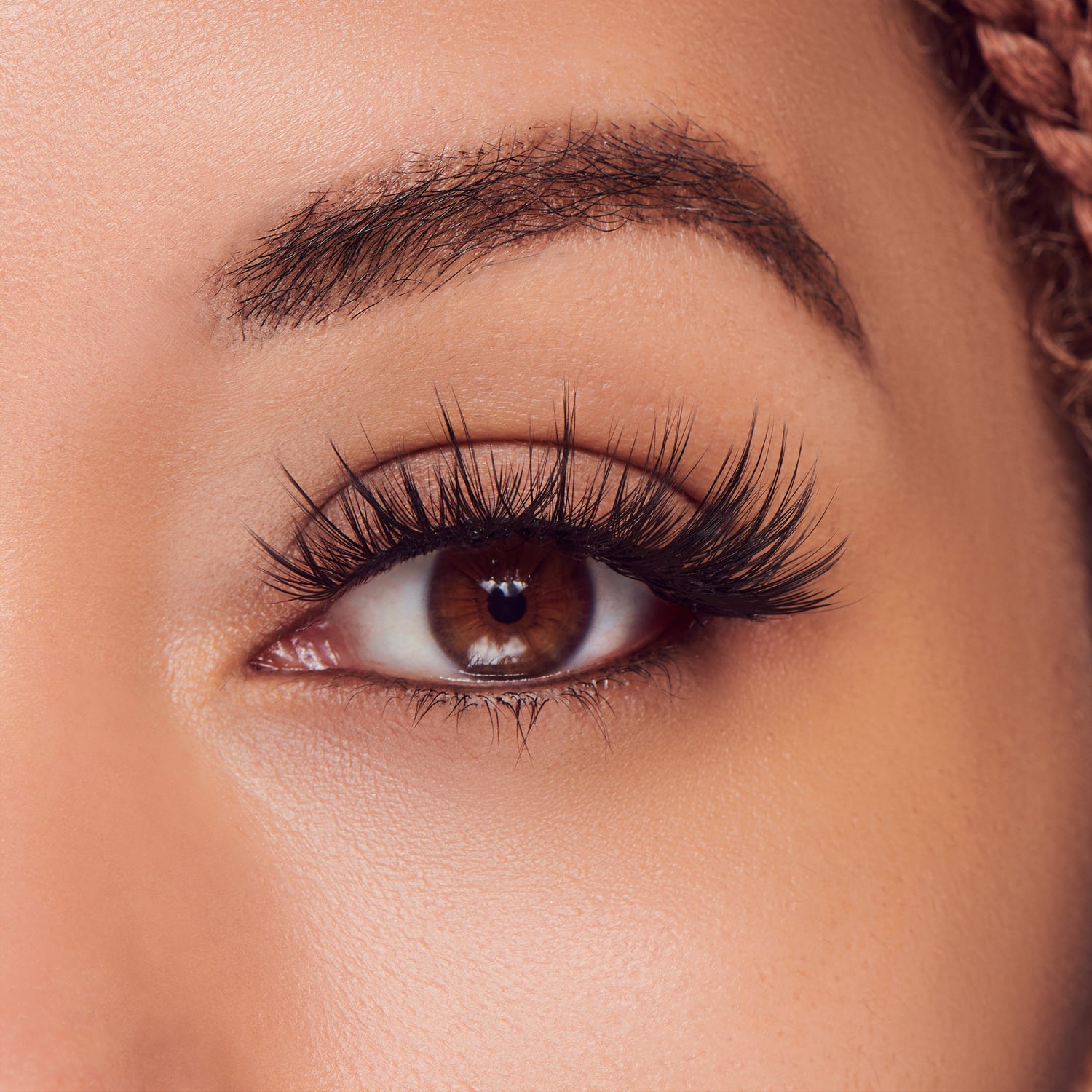 The image features a close-up of a woman's eye, showcasing her long, voluminous eyelashes and well-groomed eyebrow. The eye is a rich brown color, with a glossy appearance that highlights its depth. The skin around the eye is smooth and has a warm tone, contributing to an overall polished look. The focus on the eye emphasizes its expressive nature and the beauty of the eyelashes.