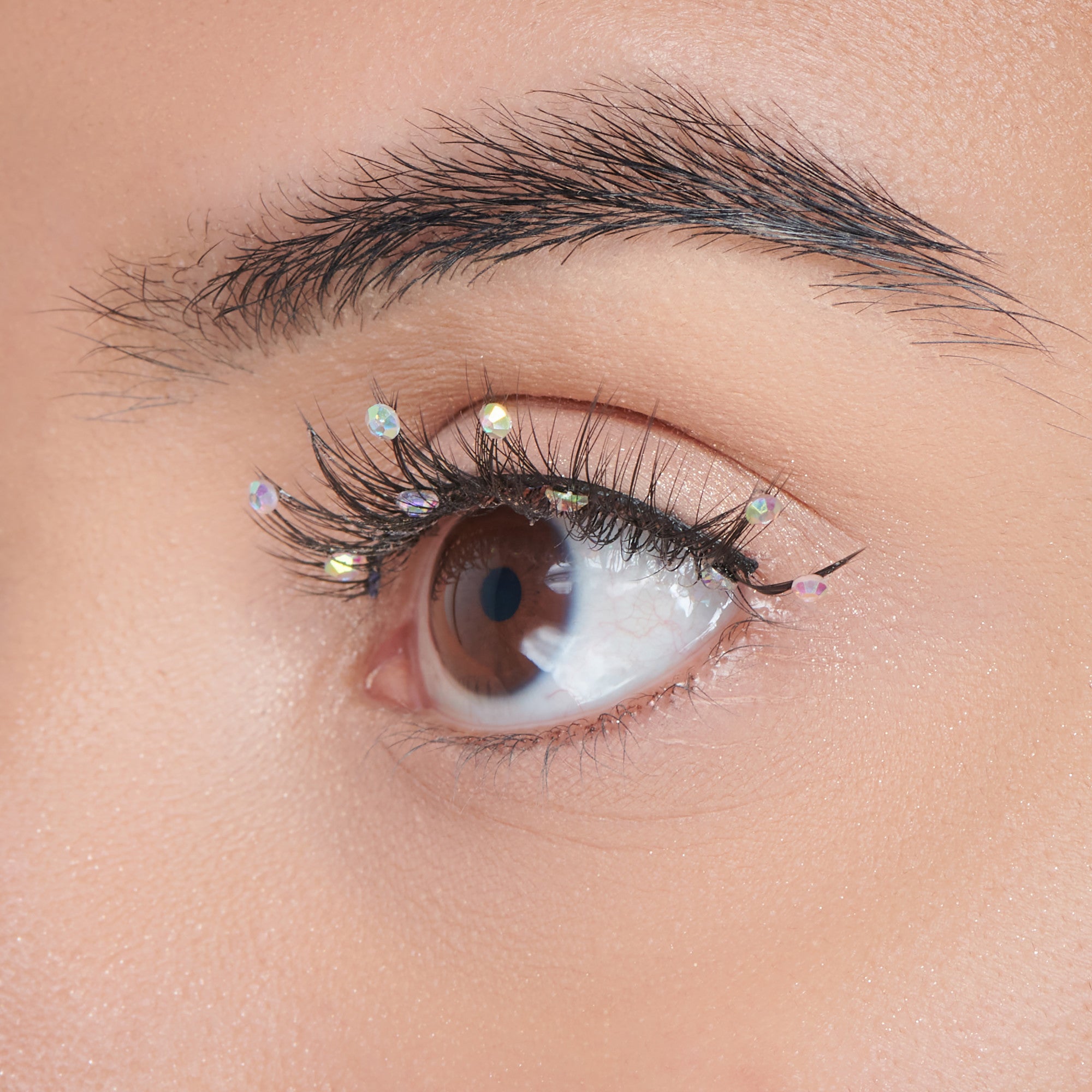 The image features a close-up of an eye with a natural makeup look. The eyelashes are adorned with small, iridescent rhinestones that add a touch of sparkle. The eyebrow is well-groomed, and the skin around the eye appears smooth and flawless. The eye itself is a warm brown color, creating a striking contrast with the decorative elements. Overall, the look is both elegant and playful, highlighting the beauty of the eye.