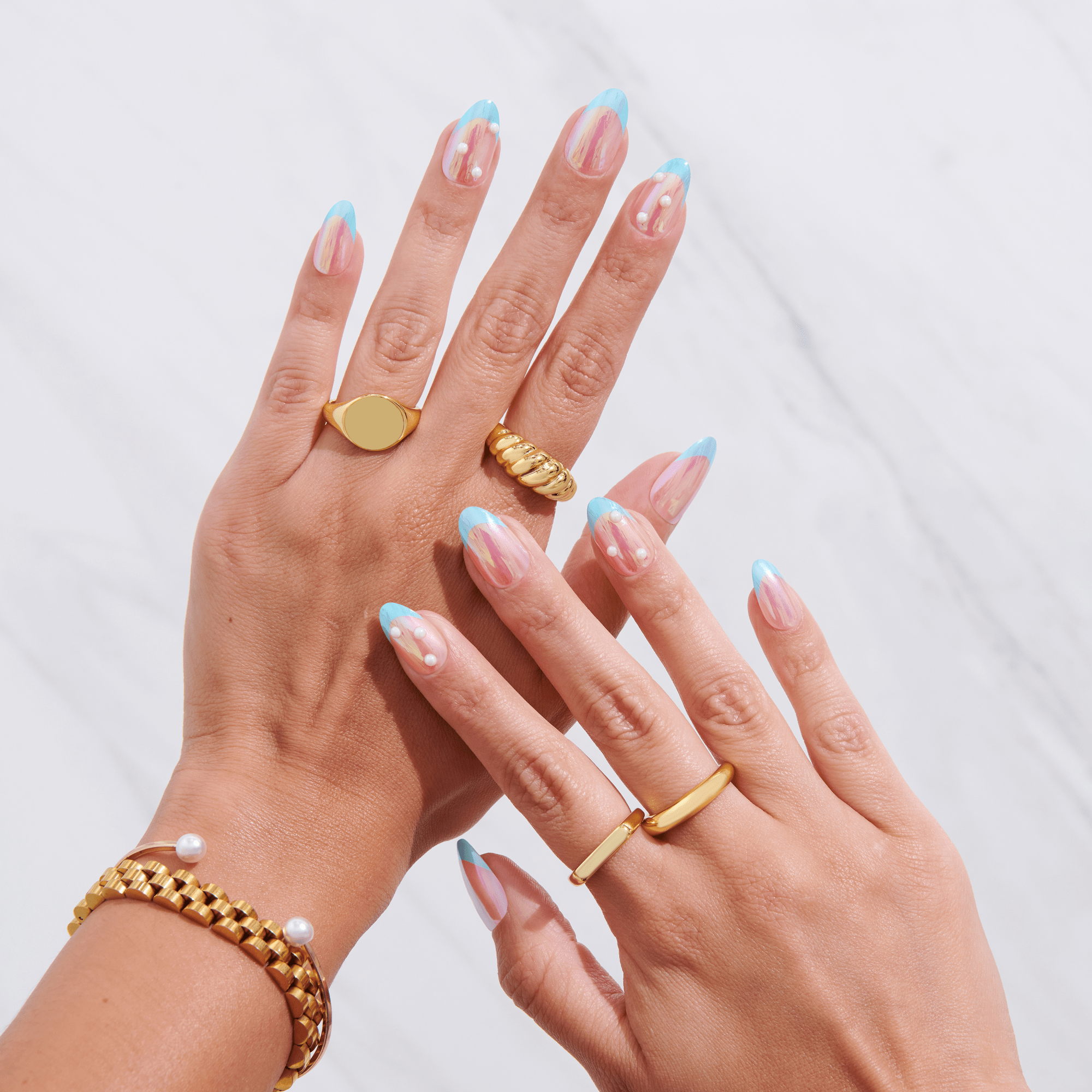 Hands with press-on nails featuring translucent pink tips with light blue French tips and white pearl accents, styled with gold rings and bracelet.