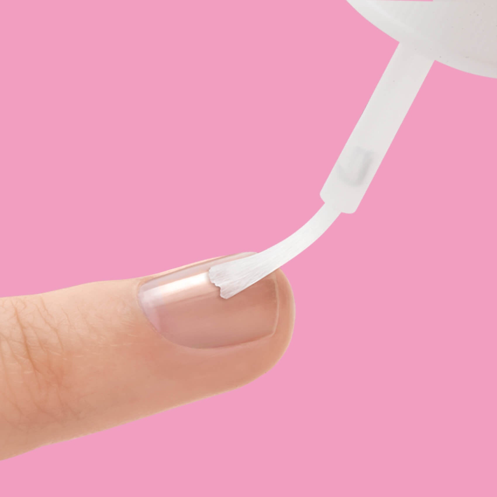 Close-up of a finger with clear nail glue being applied to the nail using a white brush applicator against a solid pink background.