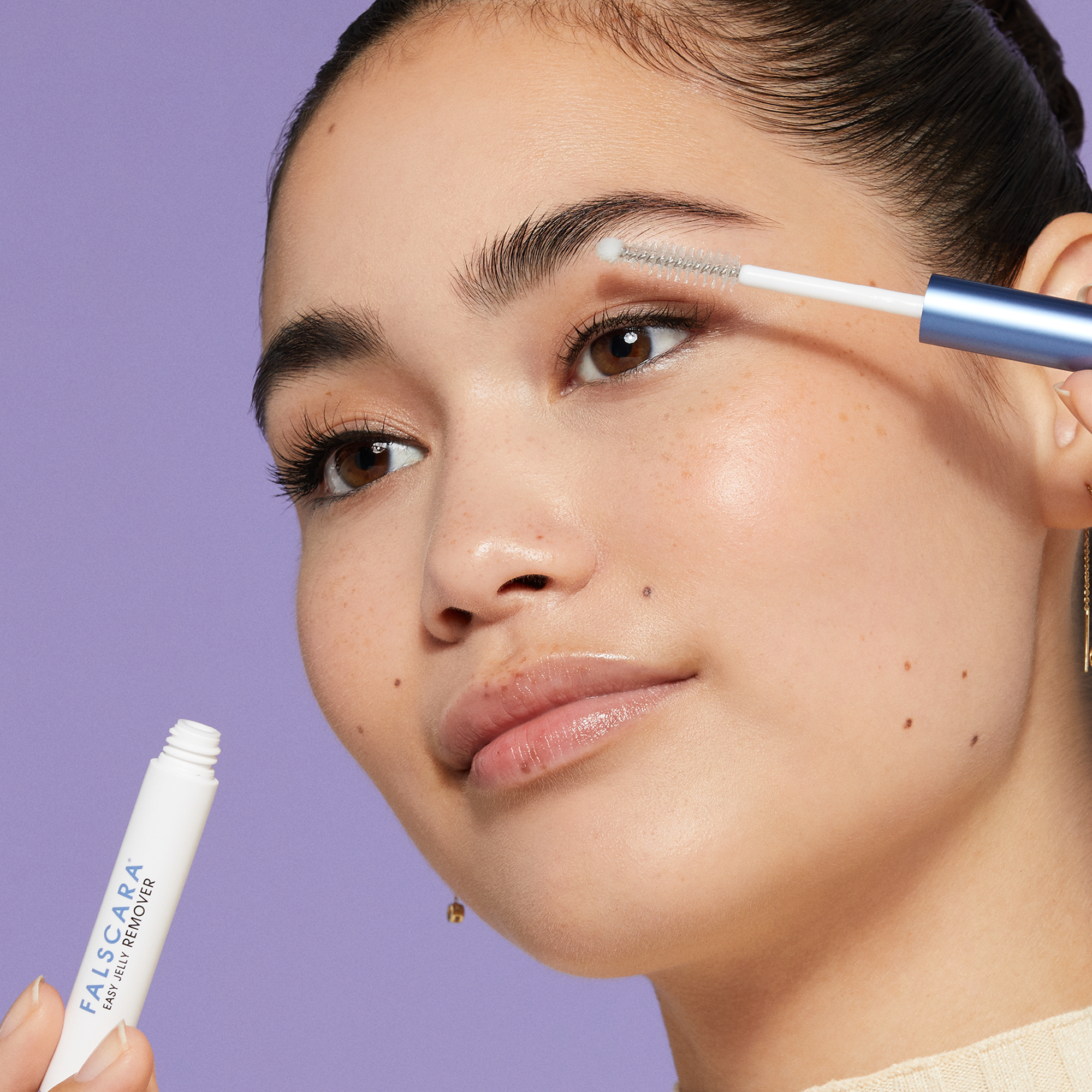 Close-up of a woman with clear skin applying a white-tube FALSCARA Easy Jelly Remover to her eyebrow using a small spoolie brush, set against a purple background.