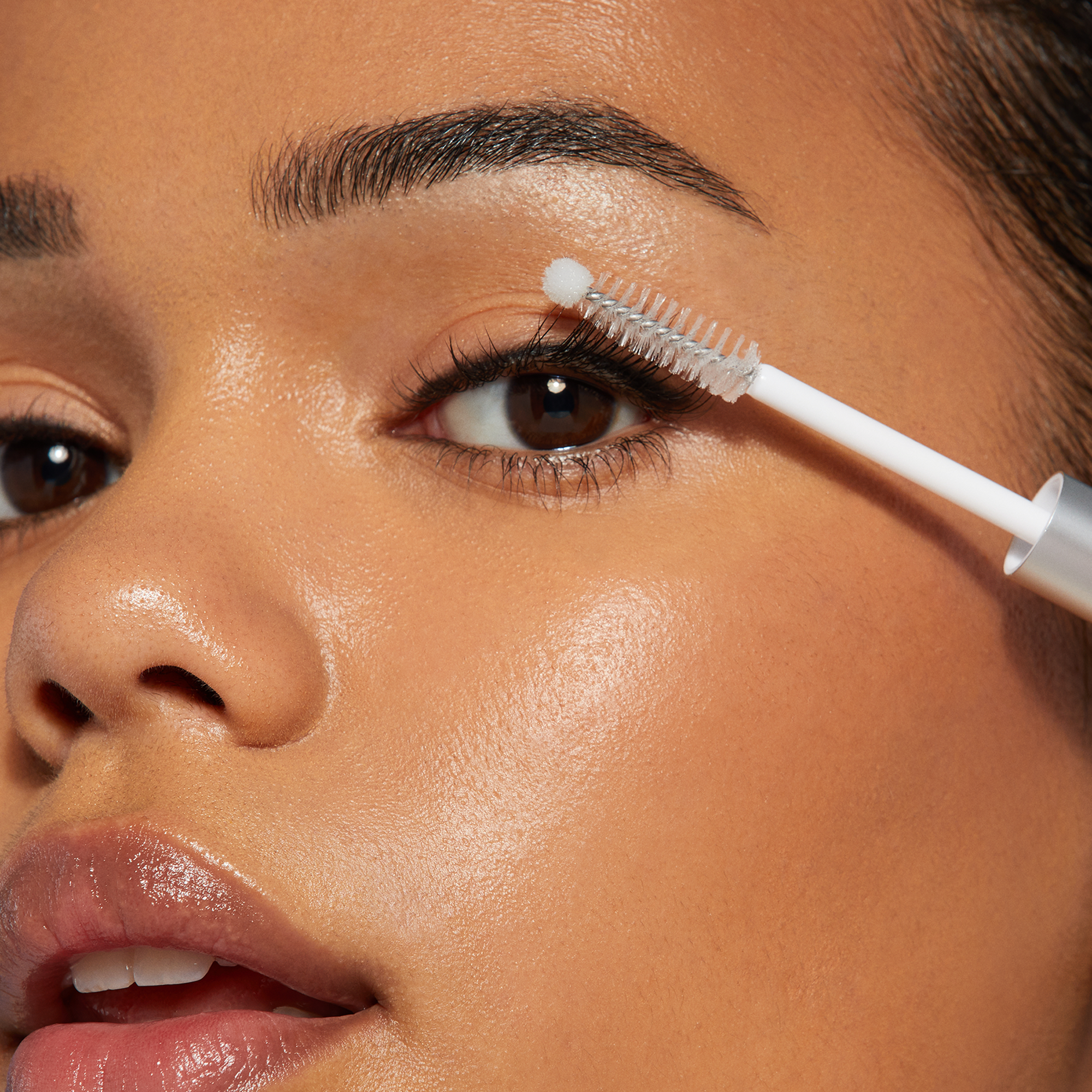 Close-up of a brown-eyed person applying clear lash adhesive with a white applicator wand featuring a small round tip near the upper eyelashes.