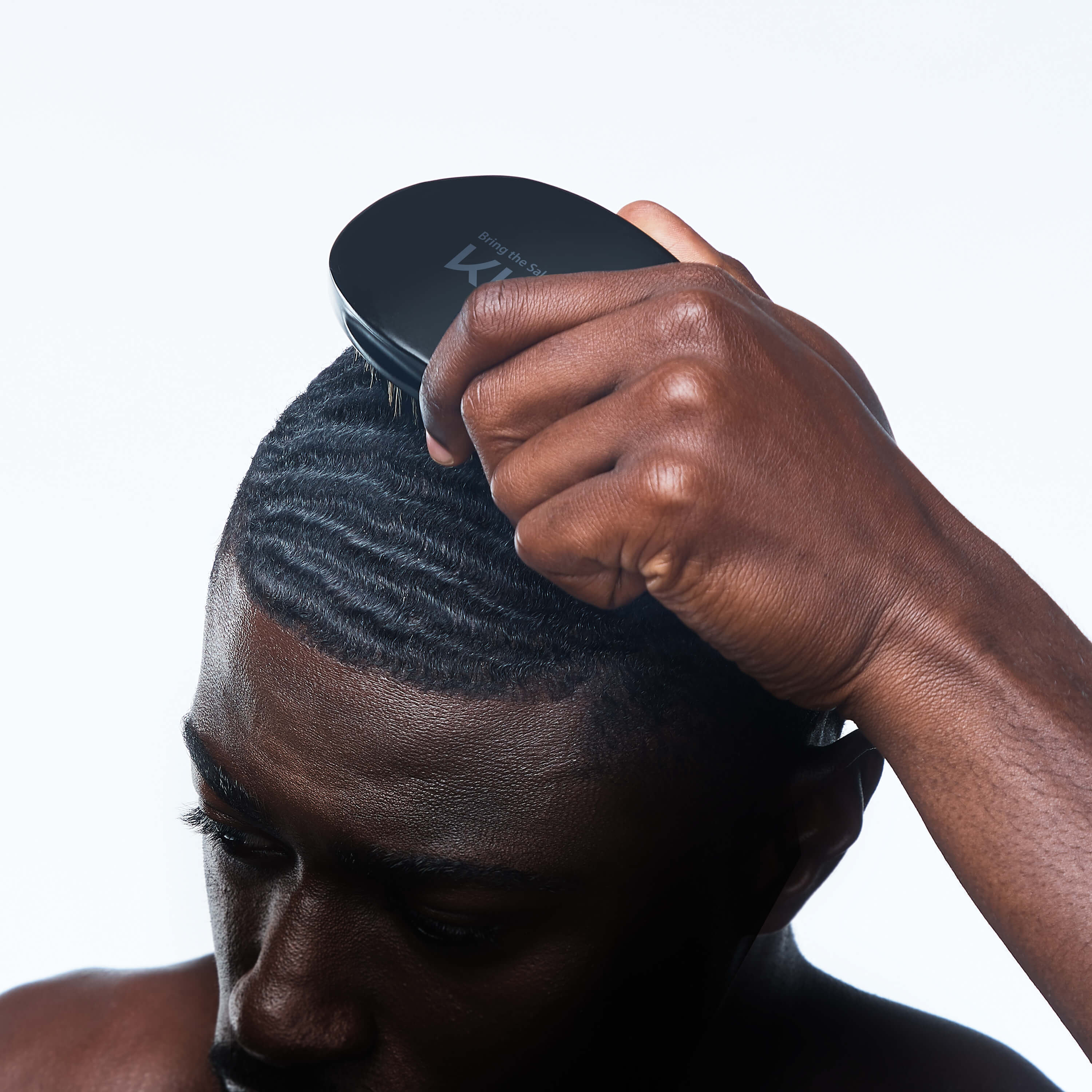 Dark-skinned man brushing short, wavy hair with a black KISS Colors & Care 360 Power Wave Premium Palm Boar Brush held in his right hand.