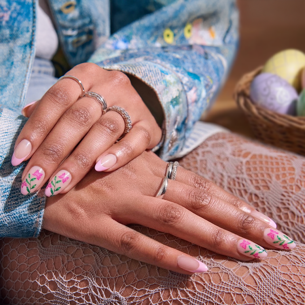 Two hands displaying pink almond nails with floral designs and silver rings.
