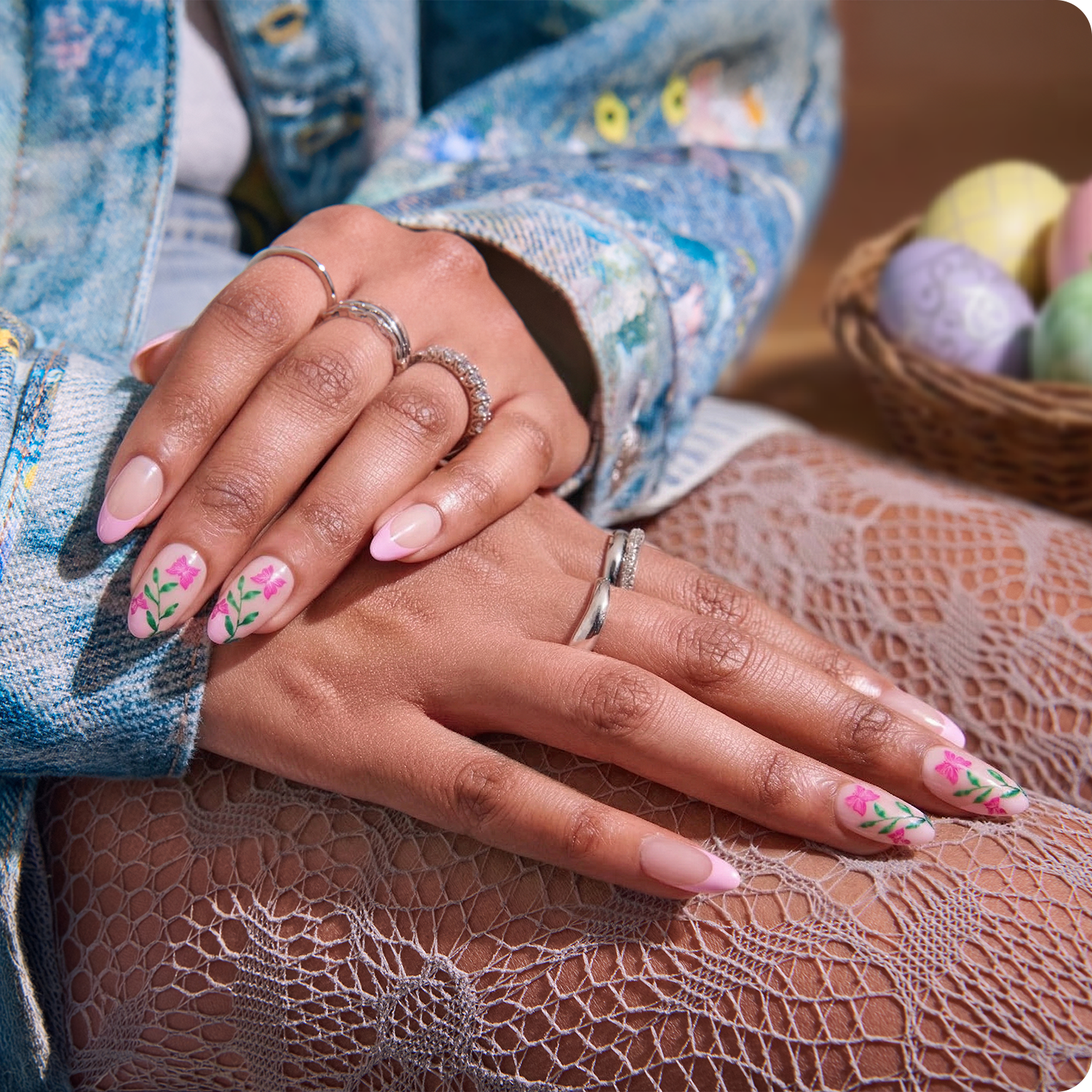 Close-up of hands with floral nail design wearing rings, with a basket of Easter eggs in the background.