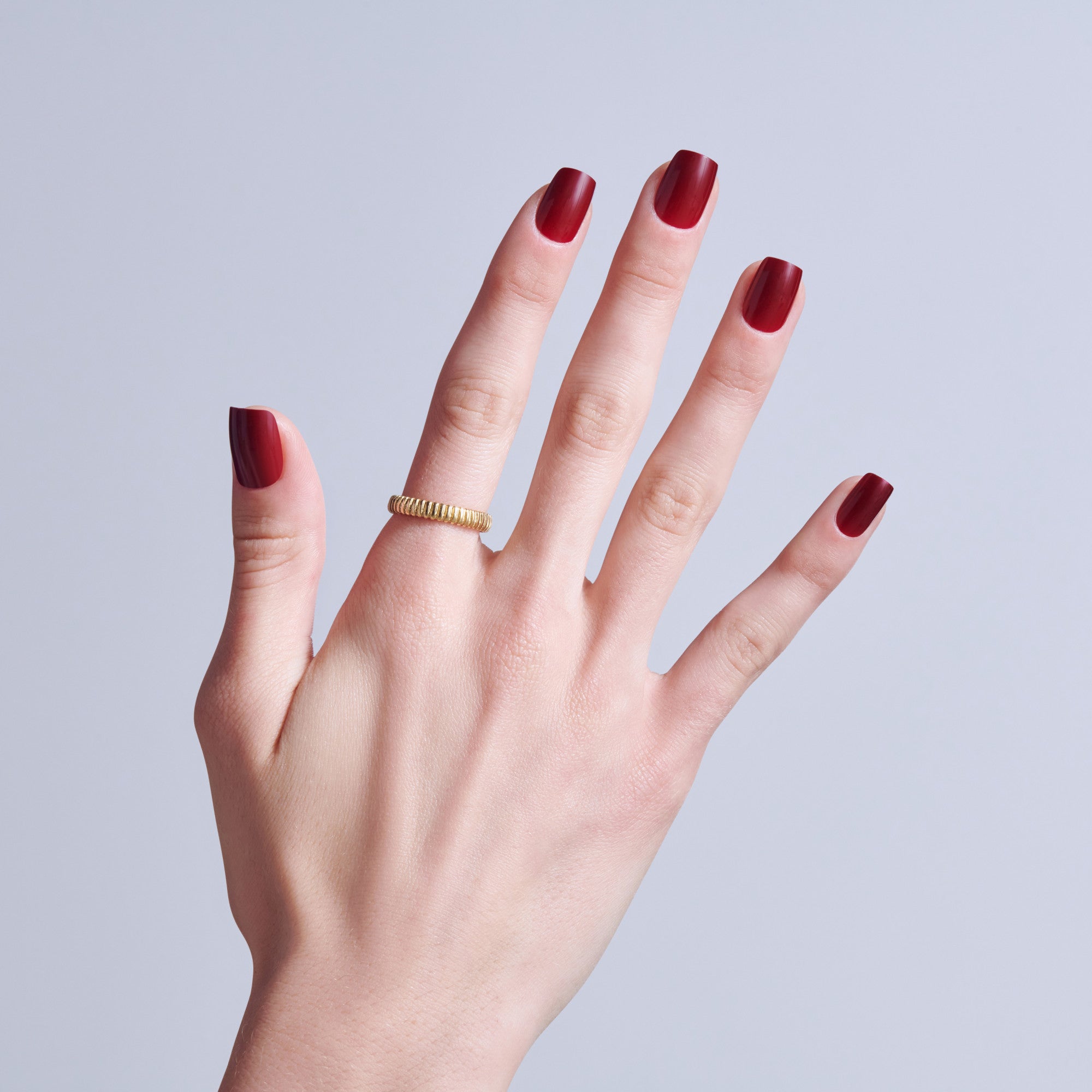 Hand displaying burgundy nail polish with gold textured ring on light background.