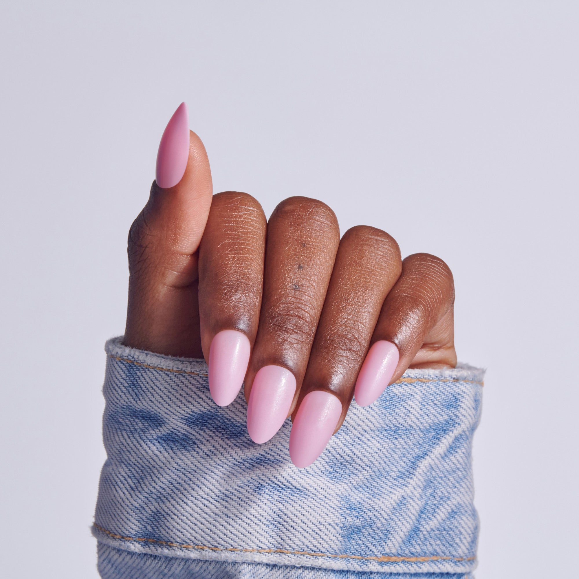 Hand with pink almond-shaped acrylic nails in light blue denim cuff against white background.