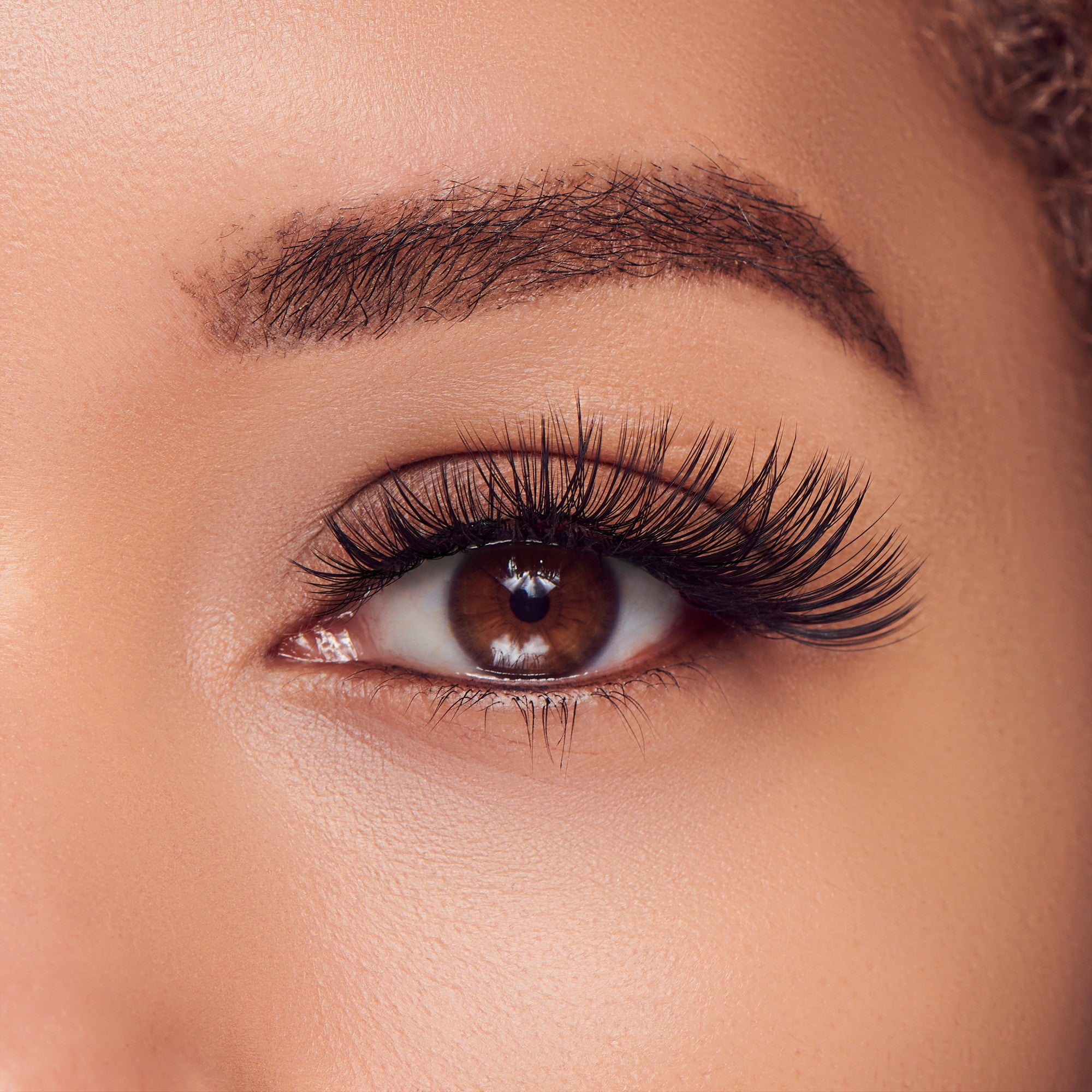 The image features a close-up of a human eye, showcasing a beautifully defined brow and long, voluminous eyelashes. The eye itself is a warm brown color, with a glossy surface that reflects light, enhancing its depth. The skin around the eye appears smooth and well-groomed, contributing to an overall polished look. The focus on the eye highlights its expressive nature and the meticulous attention to detail in the makeup, particularly the eyelashes.