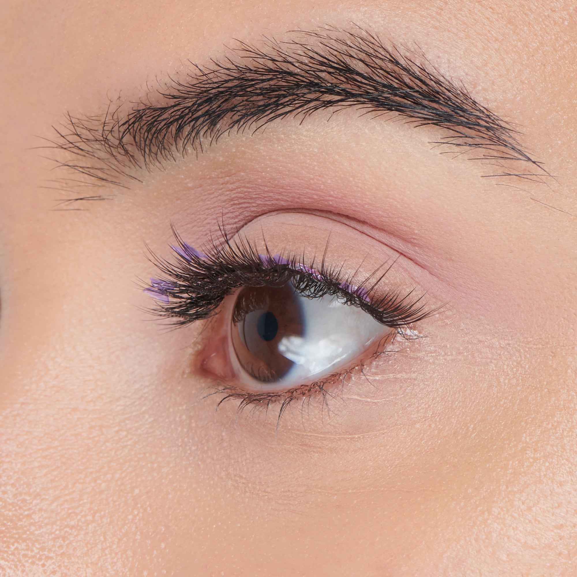 Close-up of a brown eye with long black false eyelashes featuring small purple decorative tips for a Halloween look, with soft pink eyeshadow.