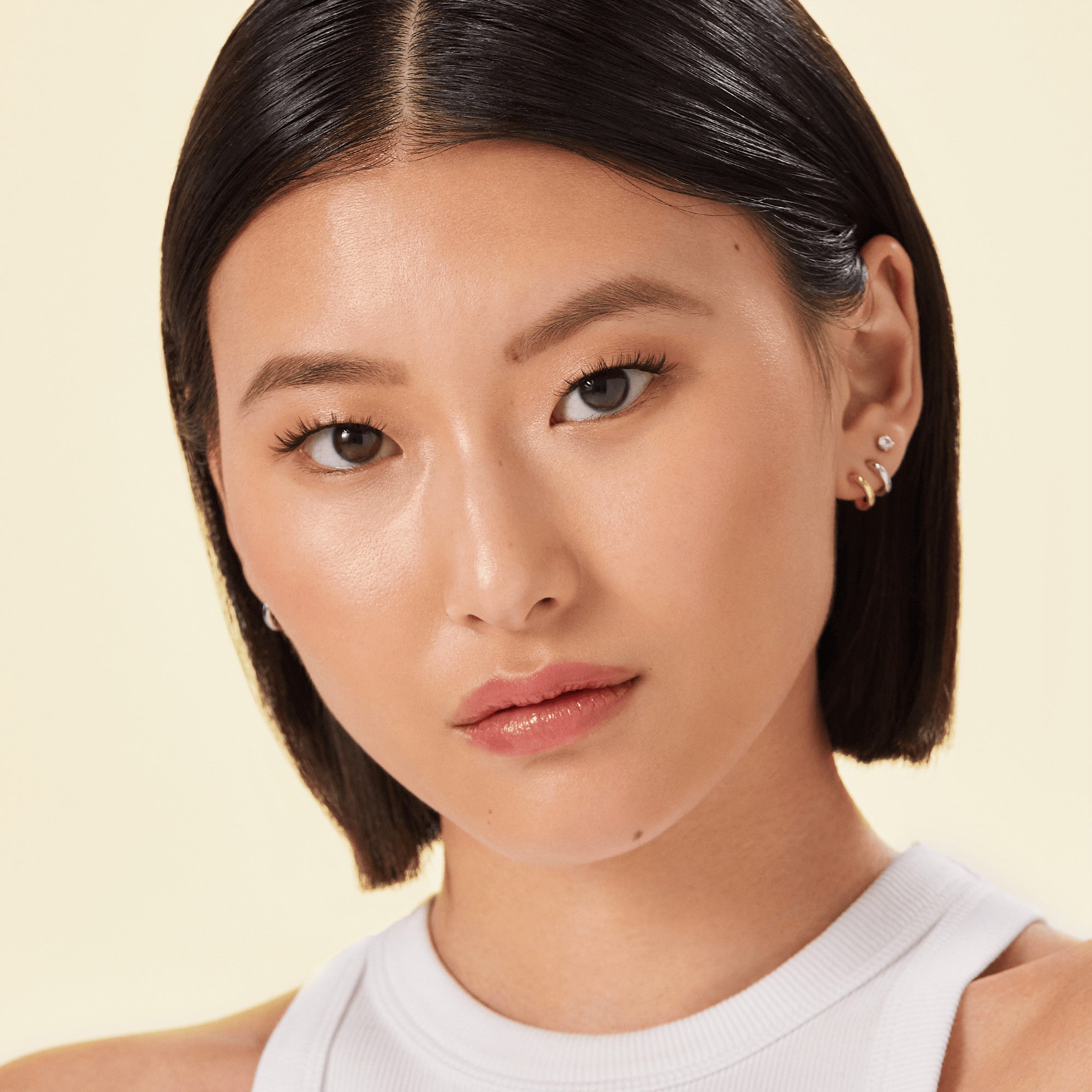 Close-up of a woman with smooth skin and modern, natural-looking false eyelashes wearing a white sleeveless top against a light background.