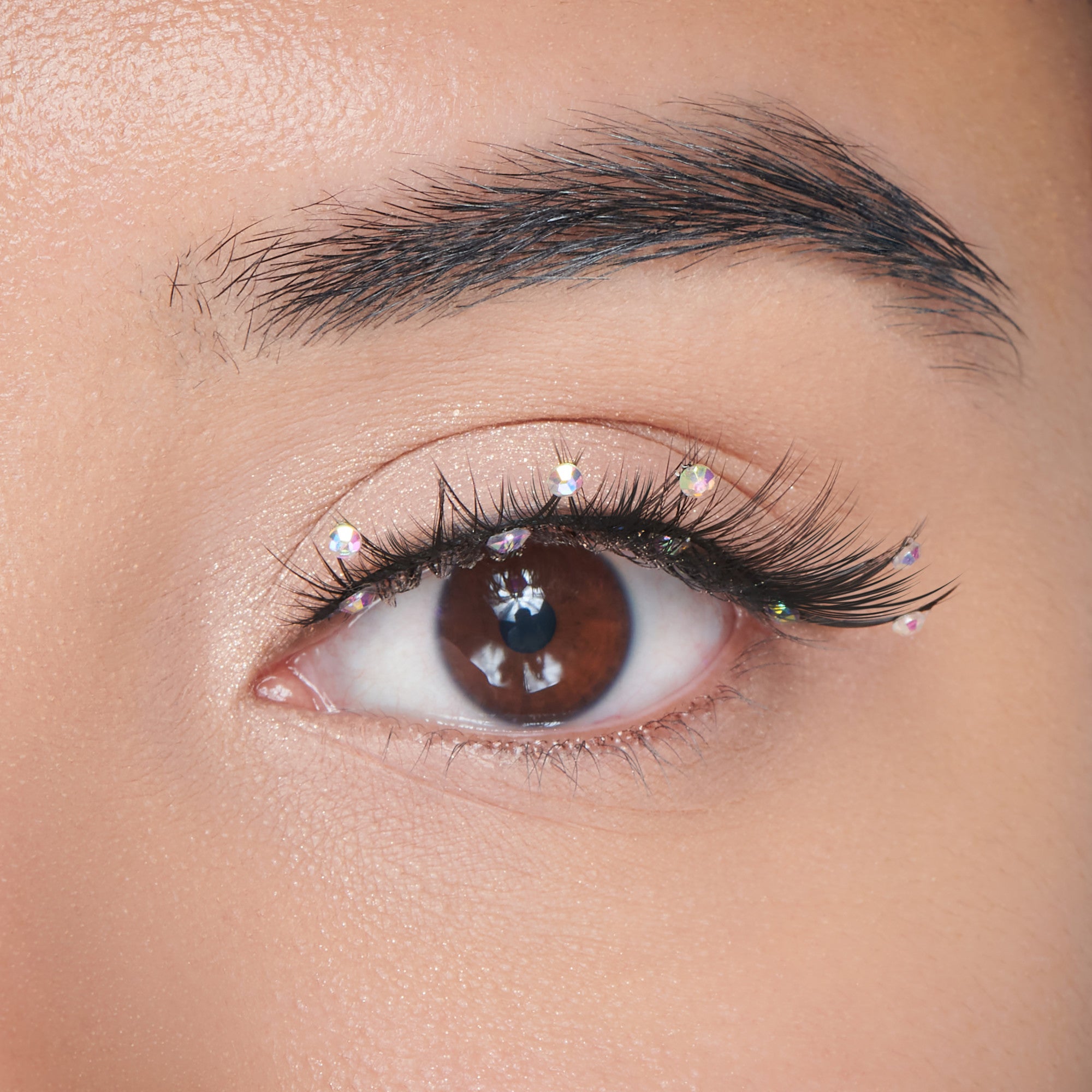 The image features a close-up of a human eye with a striking appearance. The eye has a rich brown iris and is framed by long, dark eyelashes. Delicate, sparkling embellishments are placed along the eyelid, adding a touch of glamour. The skin around the eye is smooth and has a subtle glow, enhancing the overall aesthetic. The focus on the eye creates an intimate and captivating look.