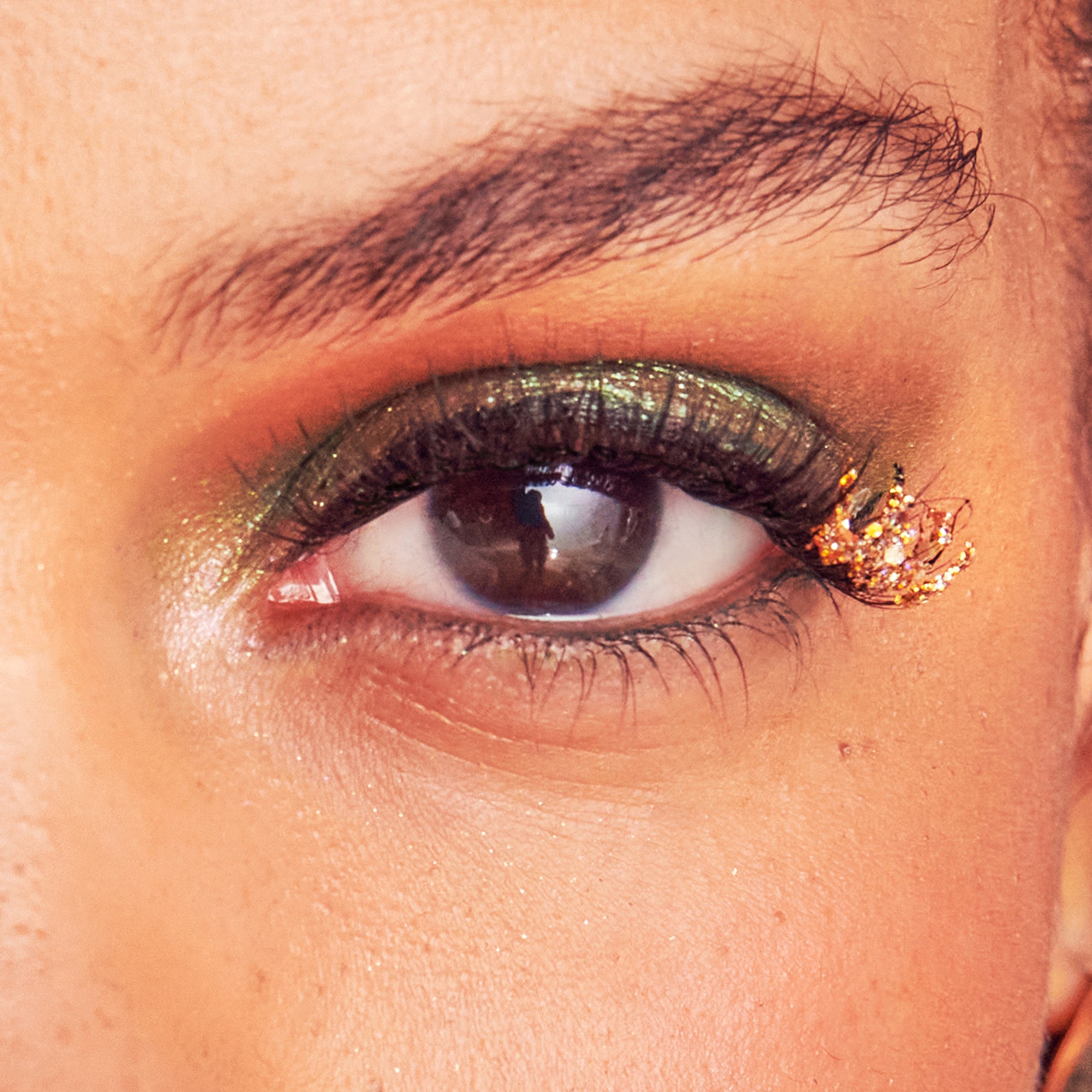 Close-up of an eye with shimmery green eyeshadow and a sparkling, gold decorative eyelash extension on the outer corner.