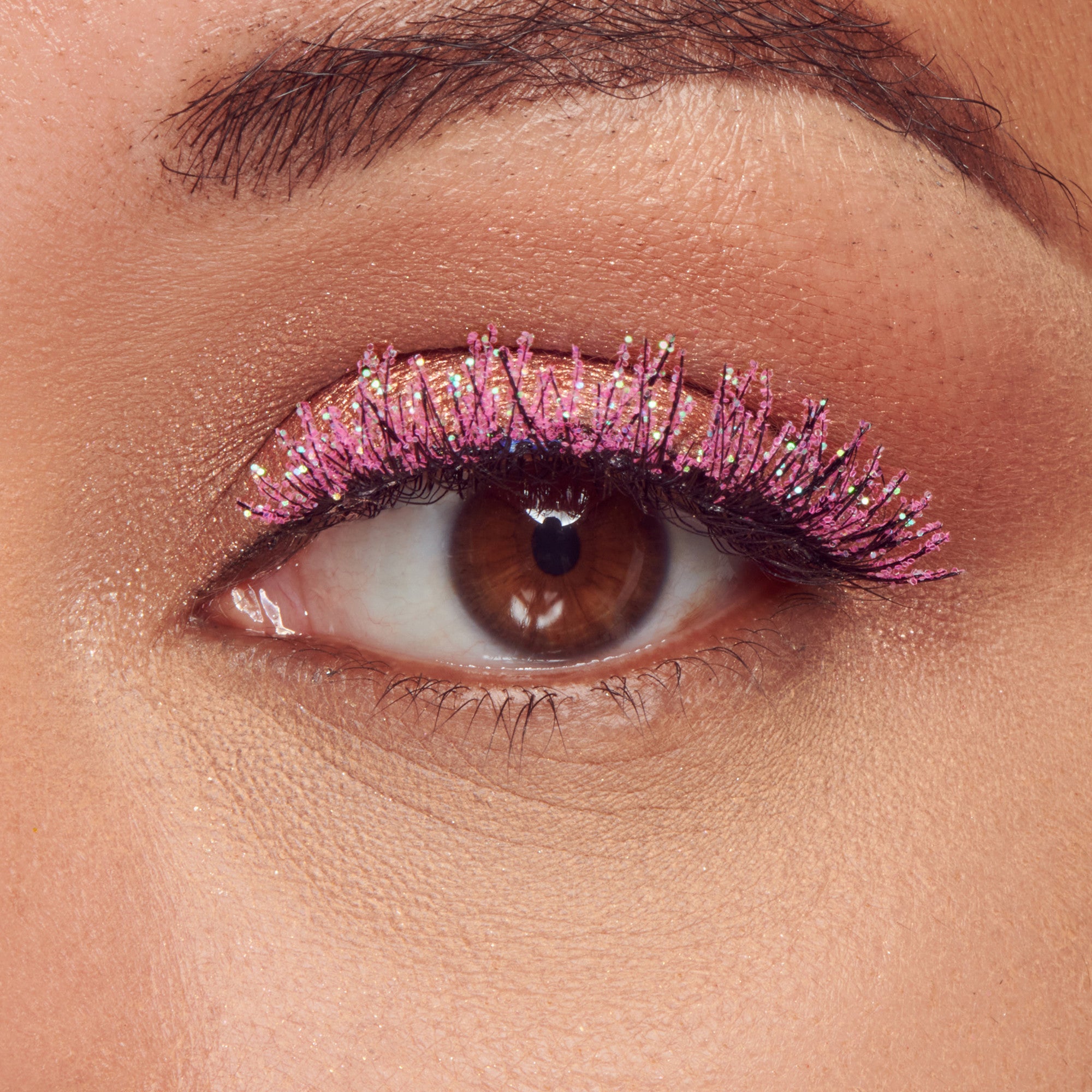 Close-up of a brown eye wearing decorated false lashes with violet glitter and multicolored gem accents, showing vibrant Halloween-themed eye makeup.