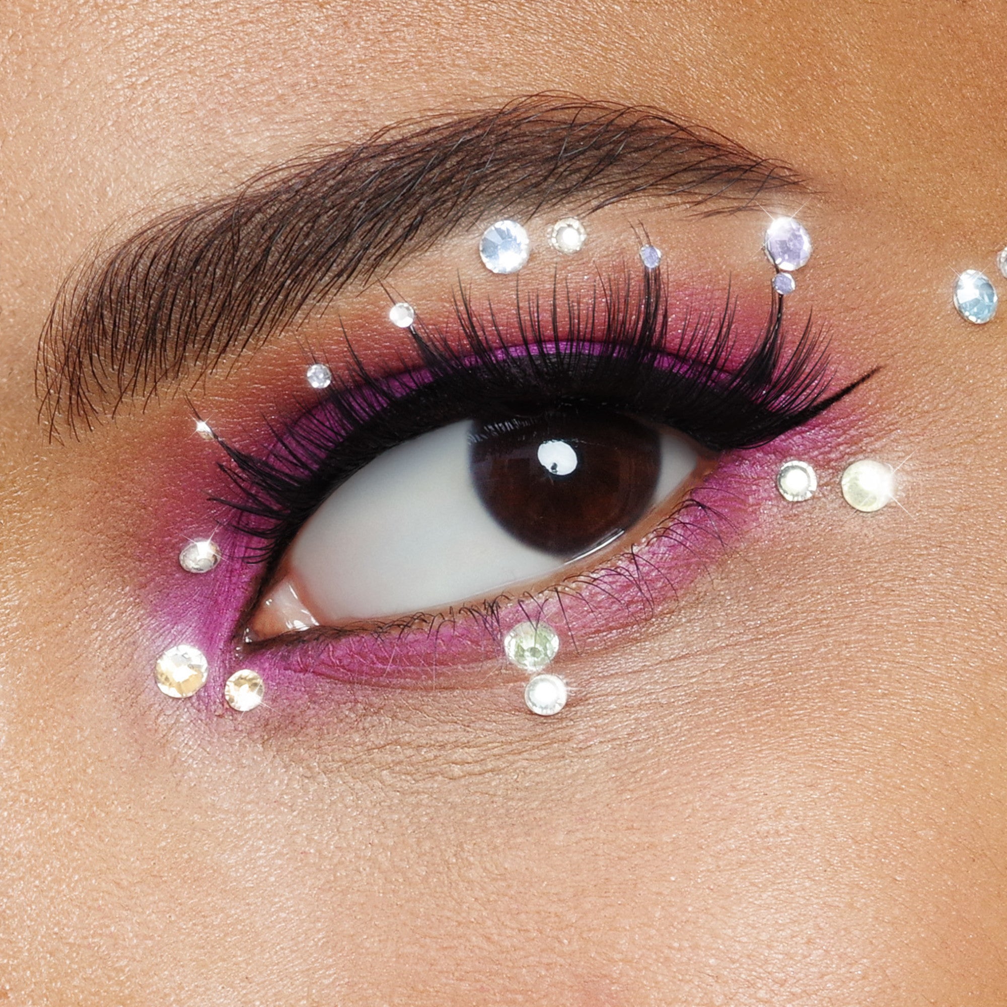 The image shows a close-up of an eye with elaborate makeup. The eyelid is adorned with vibrant pink eyeshadow, and the eyelashes are long and voluminous, likely enhanced with mascara or false lashes. Surrounding the eye, there are several small, sparkling rhinestones or gems placed strategically on the skin, adding a decorative and glamorous touch. The eyebrow is well-groomed and dark, complementing the overall bold and artistic makeup look.