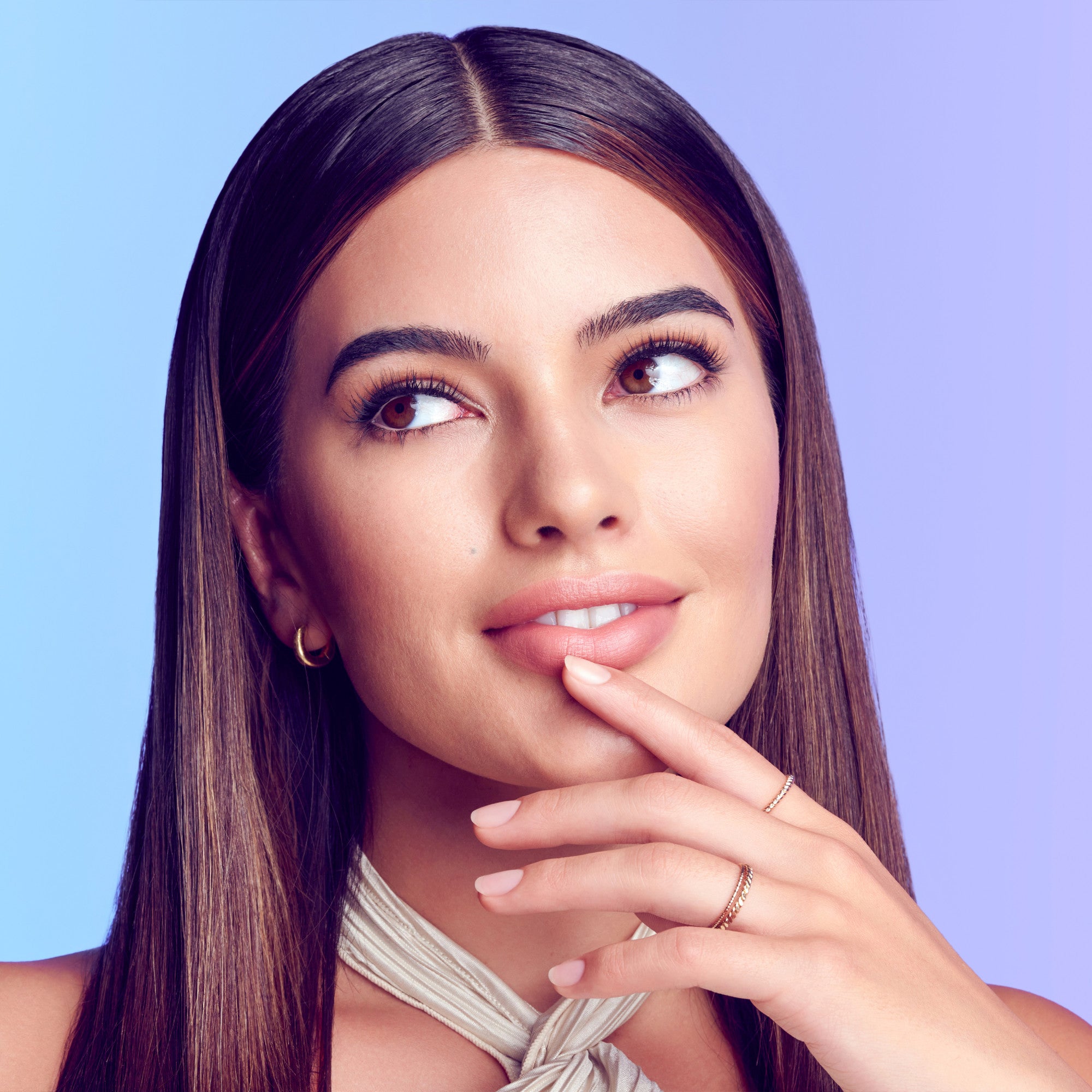 The image features a young woman with long, straight hair parted in the middle. She has a thoughtful expression, with her finger resting on her chin as she gazes slightly upward. Her makeup is natural, highlighting her features, and she wears small hoop earrings and delicate rings on her fingers. The background has a gradient of soft colors, transitioning from light blue to purple, which adds a modern and vibrant feel to the image.