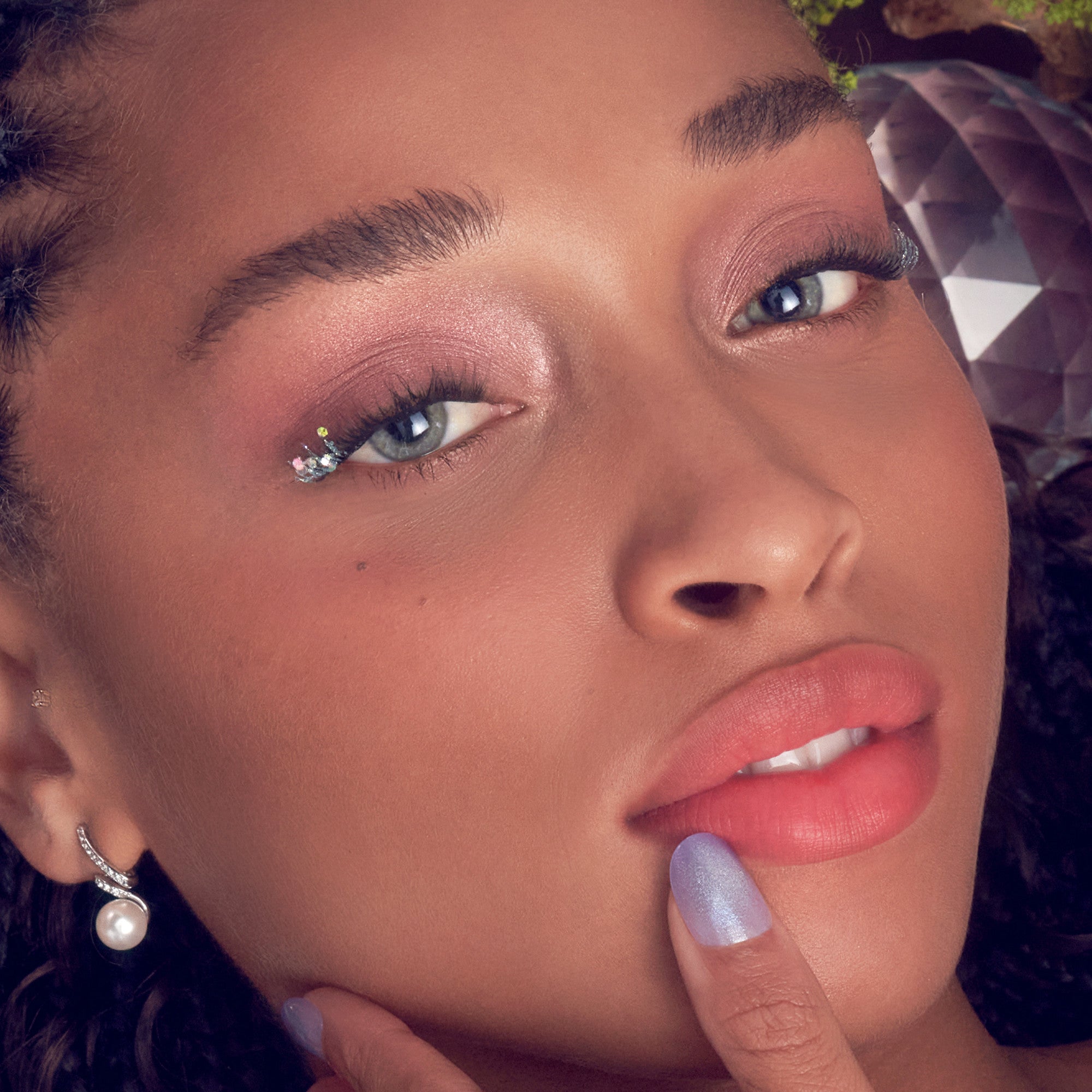 Close-up of a woman with shimmering false eyelashes featuring small crystals at the outer corners, soft pink eyeshadow, coral lips, and lavender nail polish.