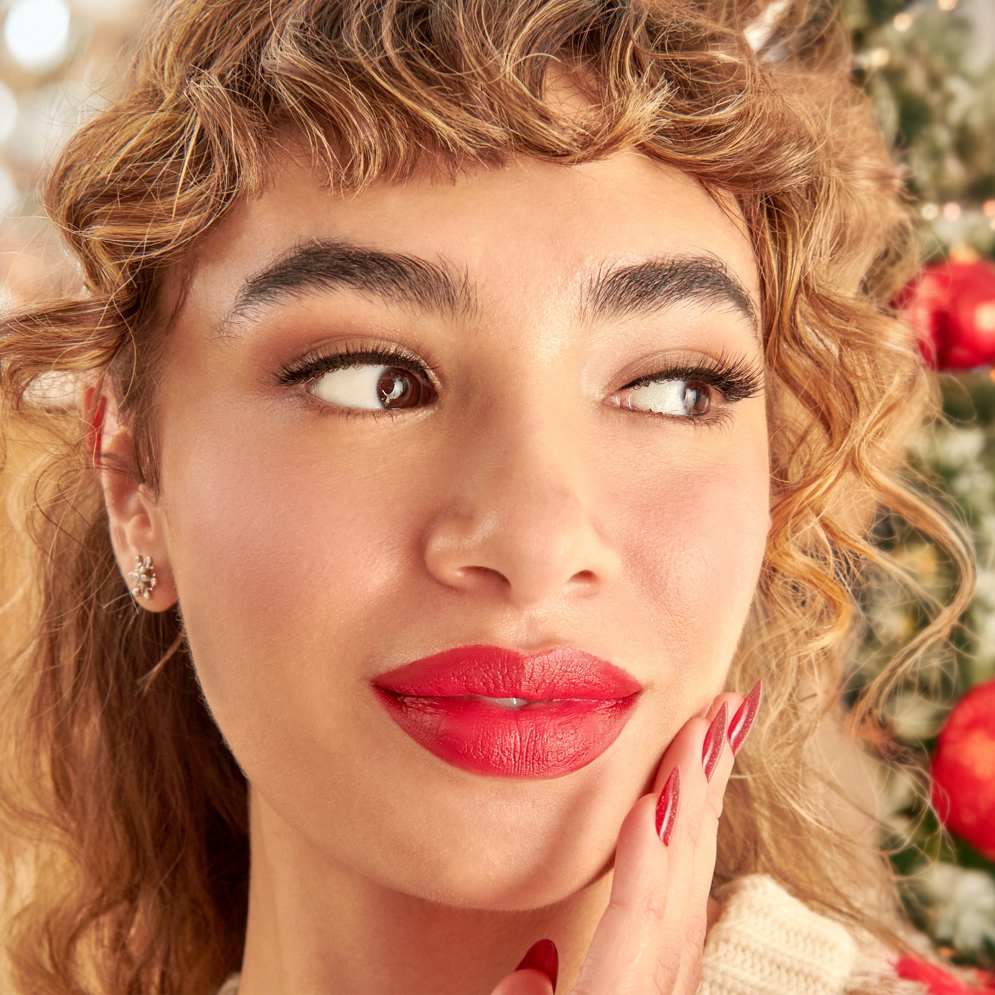 Woman with red lipstick and glittery red nails wearing natural-looking false eyelashes, posing near a decorated Christmas tree in soft lighting.